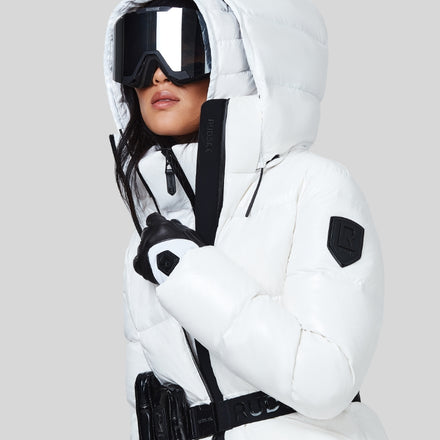 A person wearing a white puffer jacket with a hood, large black ski goggles, and black gloves, set against a plain light gray background.