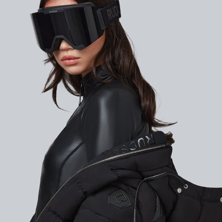 A woman wearing oversized black ski goggles, a sleek black top, and a black puffer jacket draped over her shoulder, poses against a plain light gray background.
