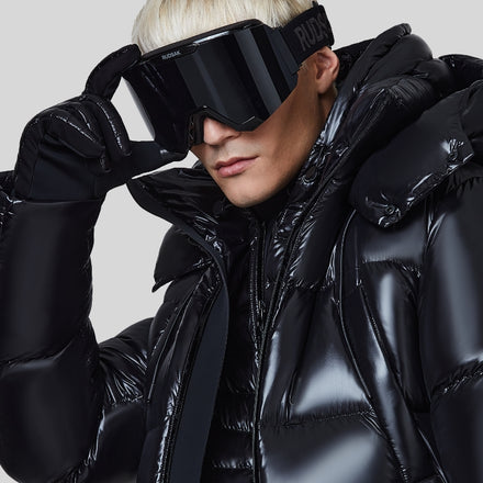 A person wearing a shiny black puffer jacket, black gloves, oversized black ski goggles, and with bleached blonde hair, poses against a plain gray background.