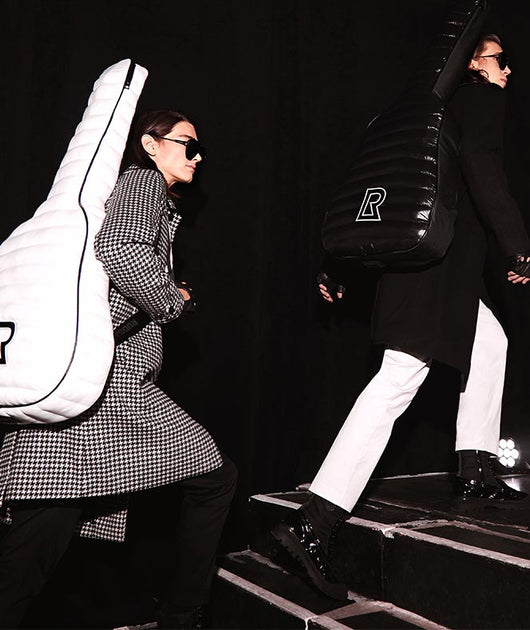 Two people wearing sunglasses and stylish coats walk up stairs, each carrying a large, padded guitar case on their backs—one case is white and the other black—against a dark background.
