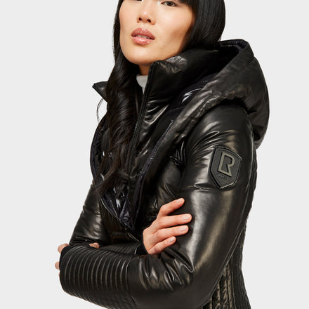 A woman with straight black hair and bangs poses in a shiny black hooded jacket with a quilted texture and an R patch on the sleeve, standing against a light gray background.