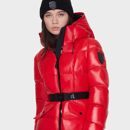 A woman wearing a shiny red puffer jacket with a hood, black beanie, and black belt stands against a plain white background. The jacket and beanie both have a black patch logo.