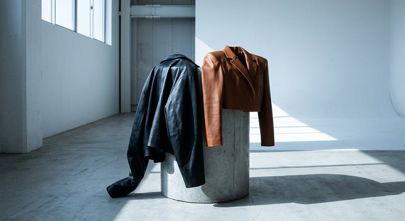 Two leather jackets, one oversized and one fitted, draped over a concrete pedestal in a minimalist studio with dramatic shadow and cool lighting.