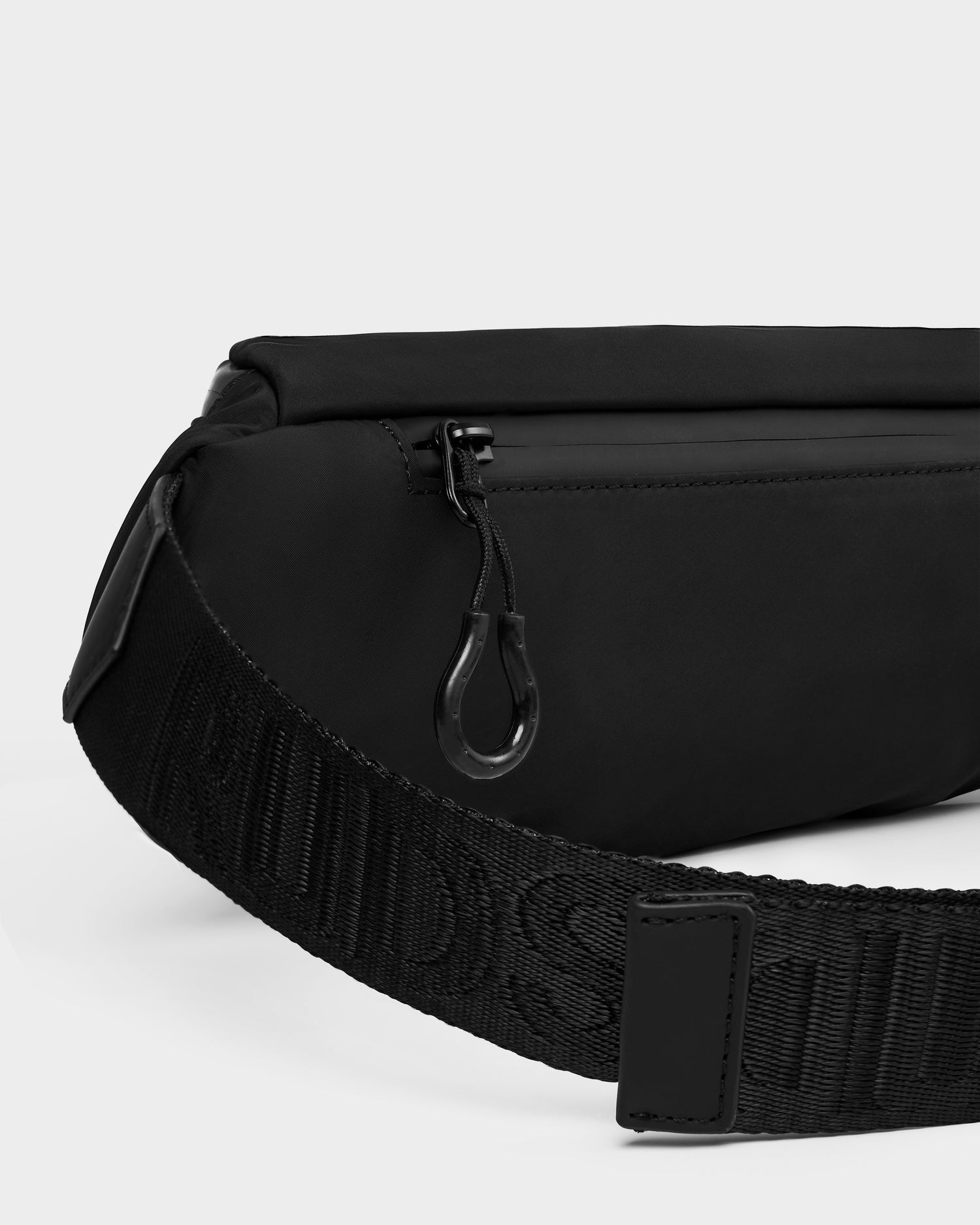 Close-up of the Rudsak RILO - 9326000 Black crossbody bag featuring a zippered pocket, wide adjustable strap with embossed text, and a black metal zipper pull.