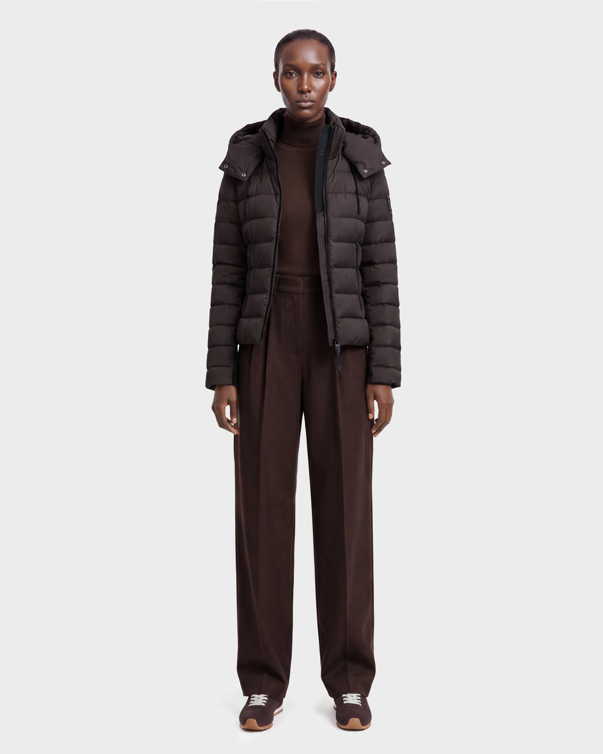 A person stands facing forward against a plain light gray background, wearing the Rudsak MELISSA - 8124824 Espresso/Black weatherproof down jacket, a dark brown turtleneck sweater, wide-leg brown pants, and sneakers.