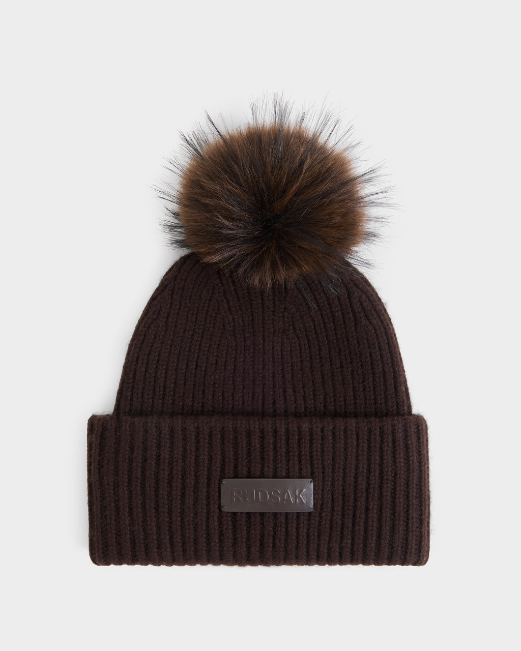 The DARA - 8425019 Espresso by Rudsak is a dark brown ribbed knit women’s winter hat, featuring a wide folded brim, a large faux fur pom-pom on top, and a rectangular leather patch with embossed branding on the front.