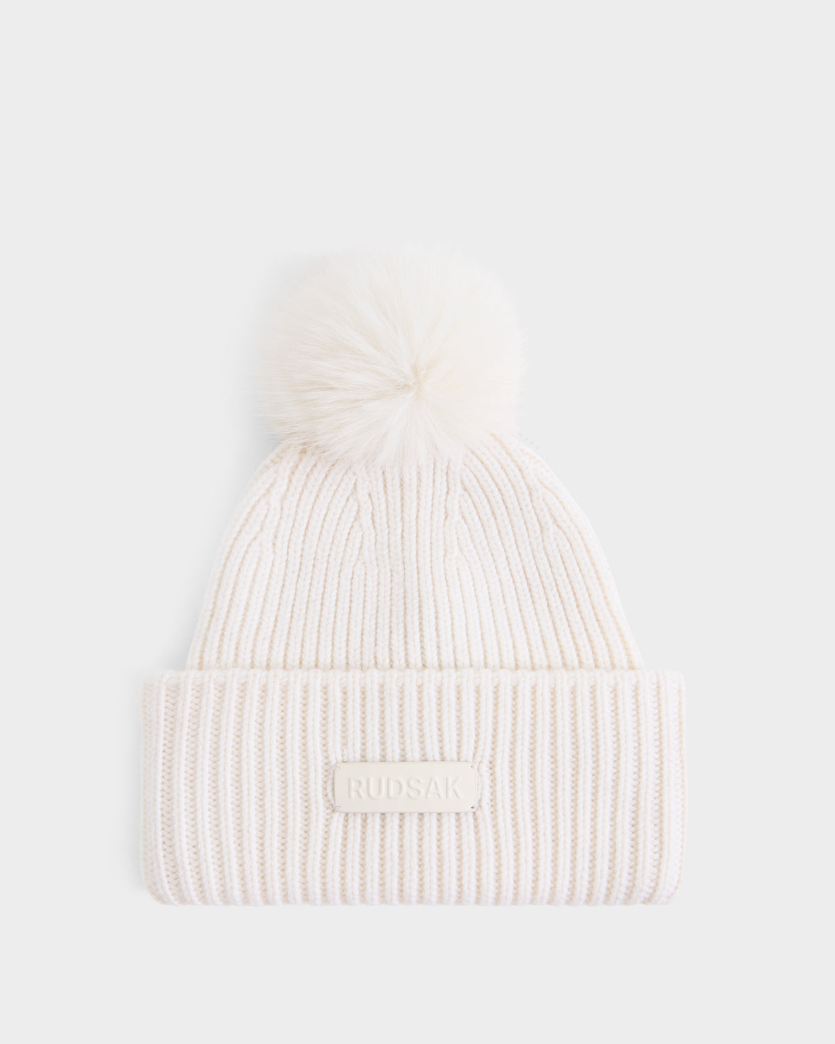 The DARA - 8425019 Vanilla by Rudsak is an off-white ribbed knit women’s tuque featuring a folded brim, a fluffy faux fur pom pom on top, and a signature RUDSAK label on the front. Crafted from a luxurious cashmere wool blend, this hat is shown against a plain white background.
