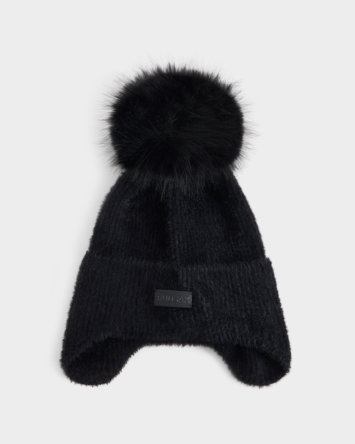 The Rudsak ICE - 8426519 Black winter hat features a black knit design with ear flaps and a large, fluffy black pom-pom on top. A small rectangular black patch accents the front of the hat, which is displayed laid flat on a light gray background.