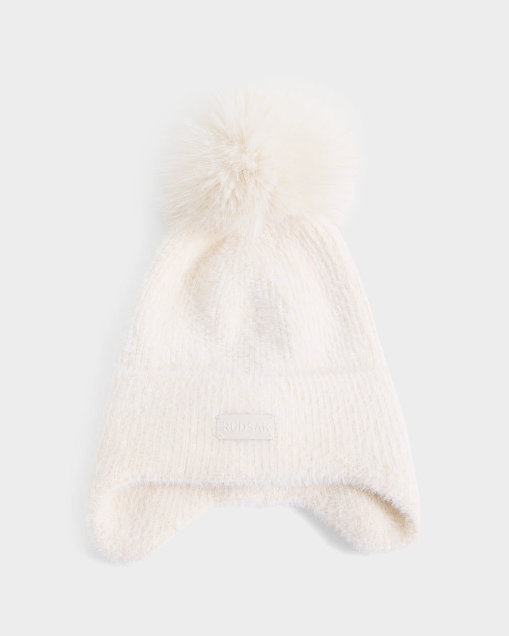 A soft white knit winter hat by Rudsak, named ICE - 8426519 Vanilla, featuring a large fluffy pom-pom on top and ear flaps. A small rectangular patch with embossed text is attached to the front brim of the hat, which is laid flat on a white background.