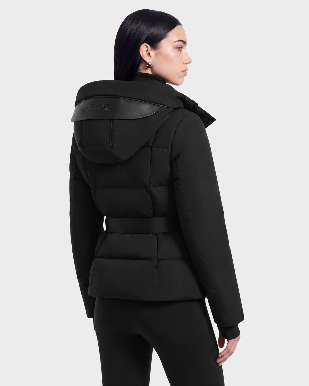 A woman with straight dark hair is wearing the Rudsak ADELINE - 8124544 black belted women’s down puffer jacket and black pants, standing in a three-quarter view facing left against a plain light background.