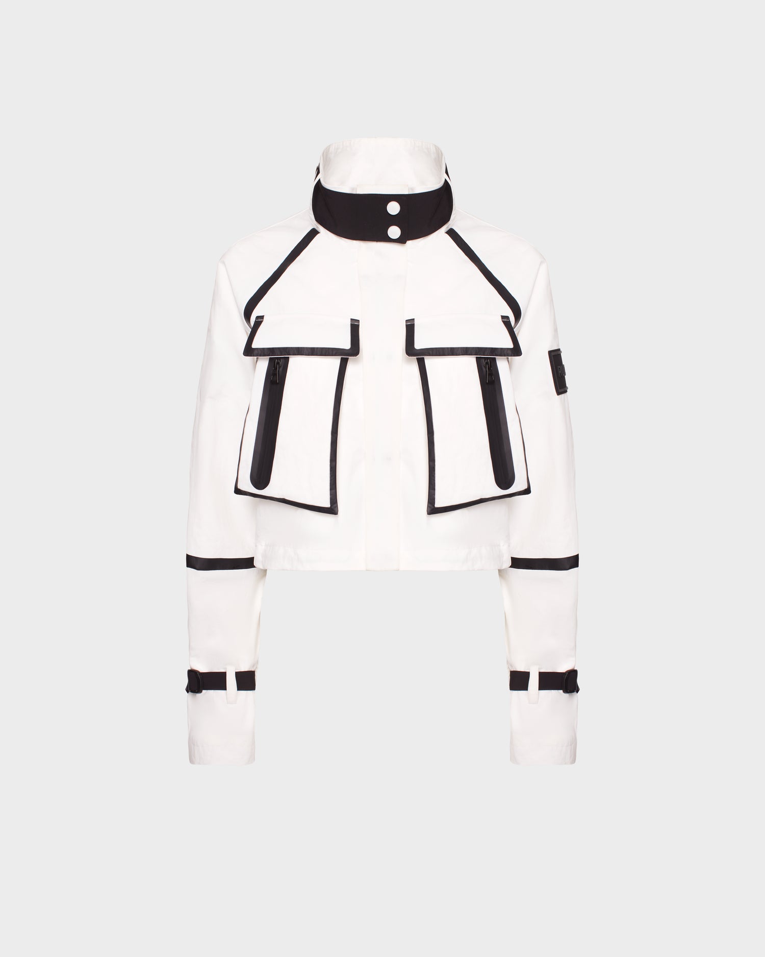 The ALODIE - 8124043 Cotton White by Rudsak is a stylish women's cropped jacket in white with black trim, large flap pockets, zippered accents, high buttoned collar, and adjustable sleeve straps. Shown on a light gray background.
