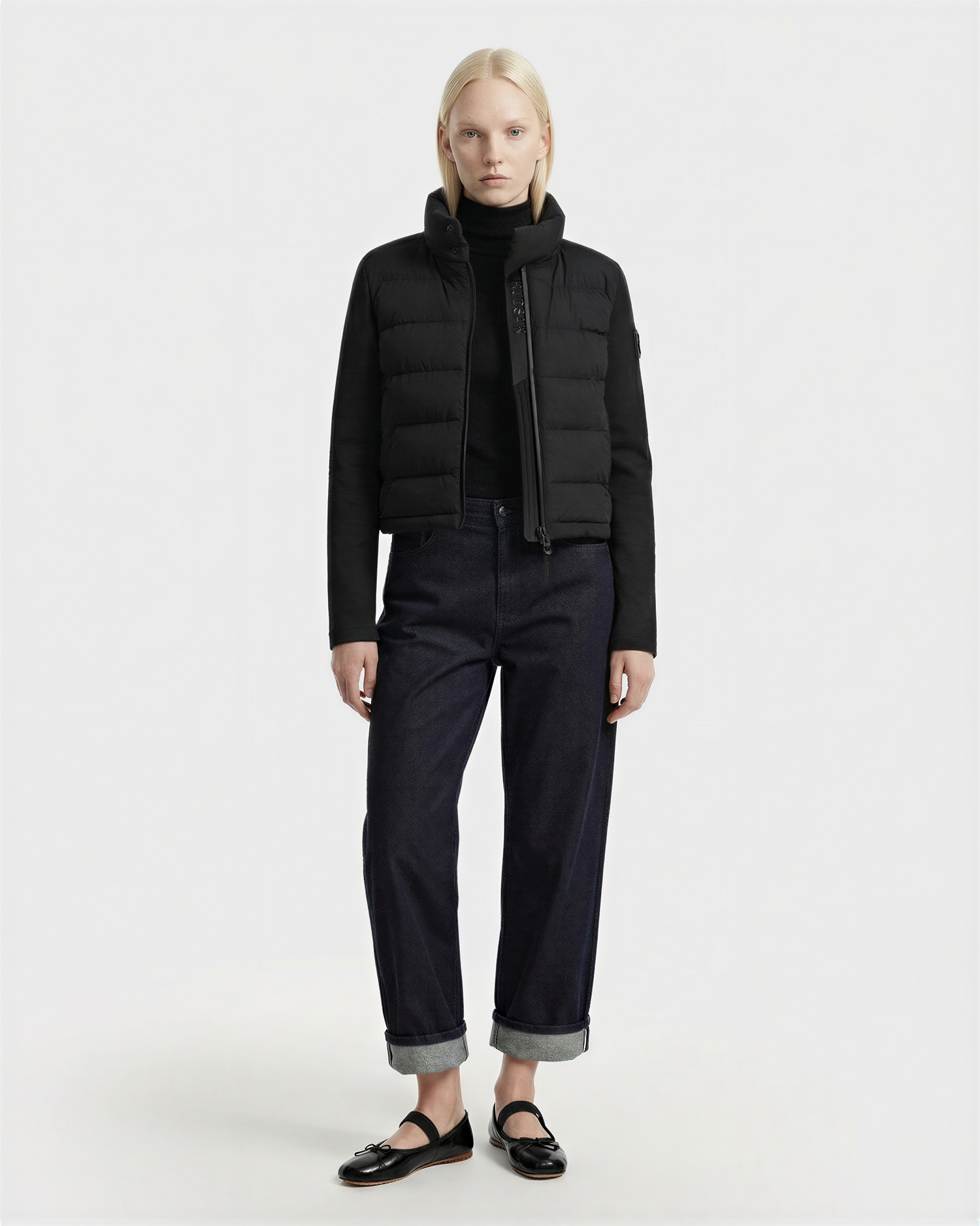 A person with long blonde hair stands against a plain background, wearing the Rudsak AUSHA - 8125026 Black women’s cropped down jacket featuring RDS-certified down, paired with a black turtleneck, dark cuffed jeans, and black loafers.