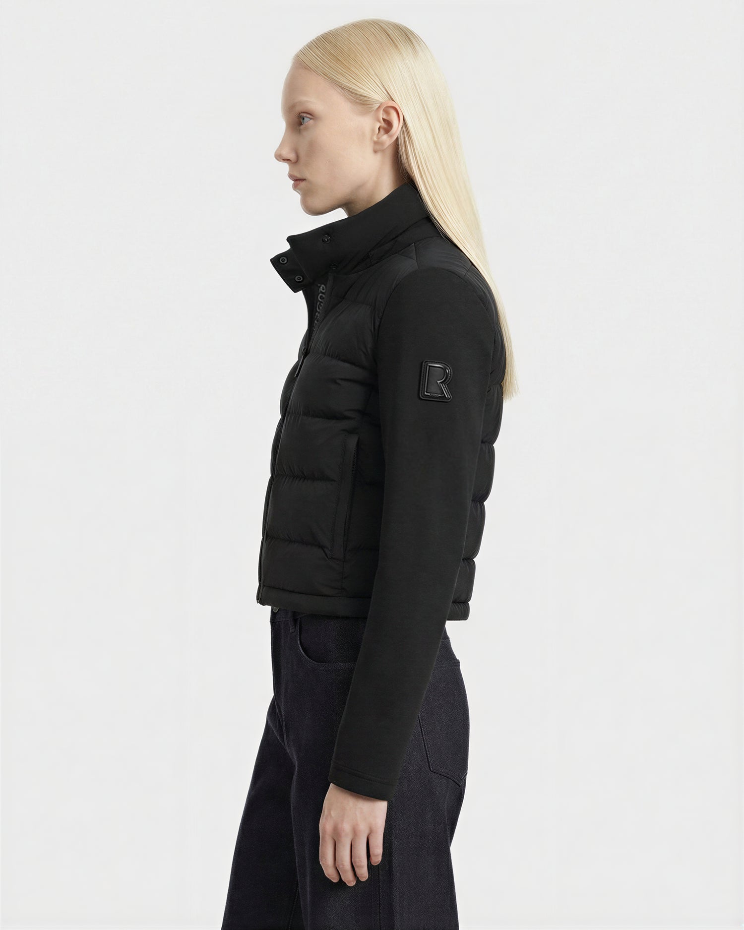 A person with straight, long blonde hair is shown in profile wearing the Rudsak AUSHA - 8125026 Black RDS-certified down puffer jacket with a high collar and sleeve patch, paired with dark pants, against a plain white background.