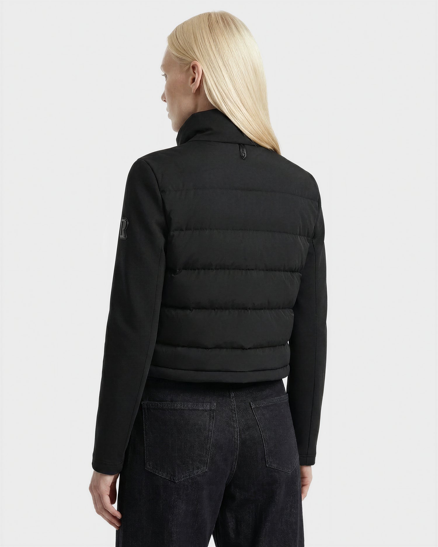 A person with long, straight blonde hair wears the Rudsak AUSHA - 8125026 Black women’s cropped down jacket and dark jeans, standing with their back to the camera against a plain, light background.