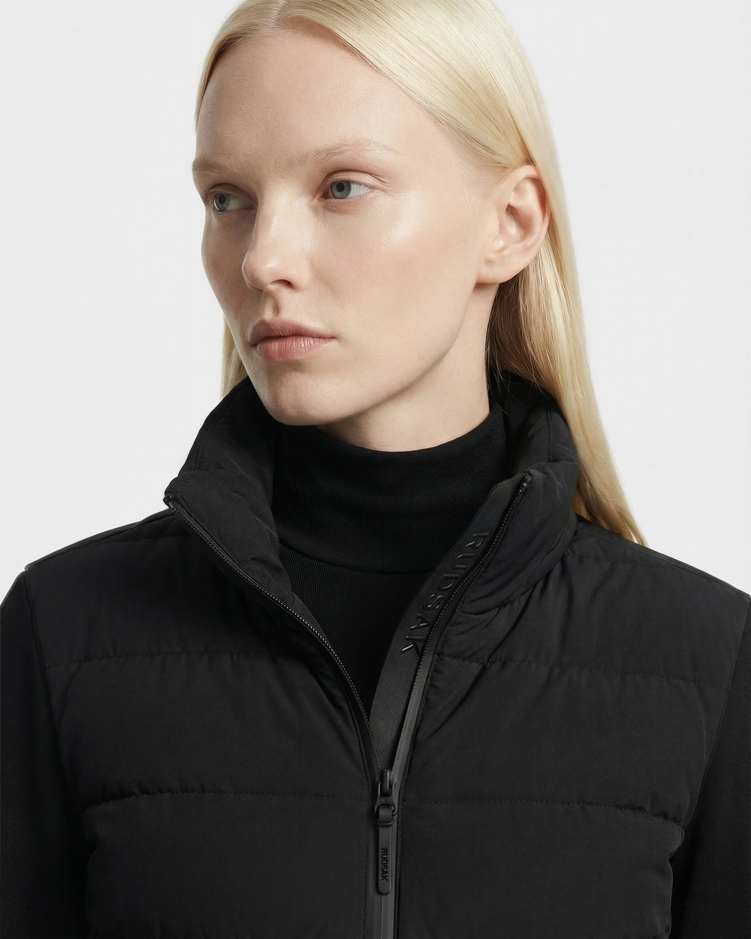 A woman with straight, platinum blonde hair wears a black turtleneck and the Rudsak AUSHA - 8125026 Black lightweight down jacket with a high collar. She looks to the side with a neutral expression against a plain, light background.