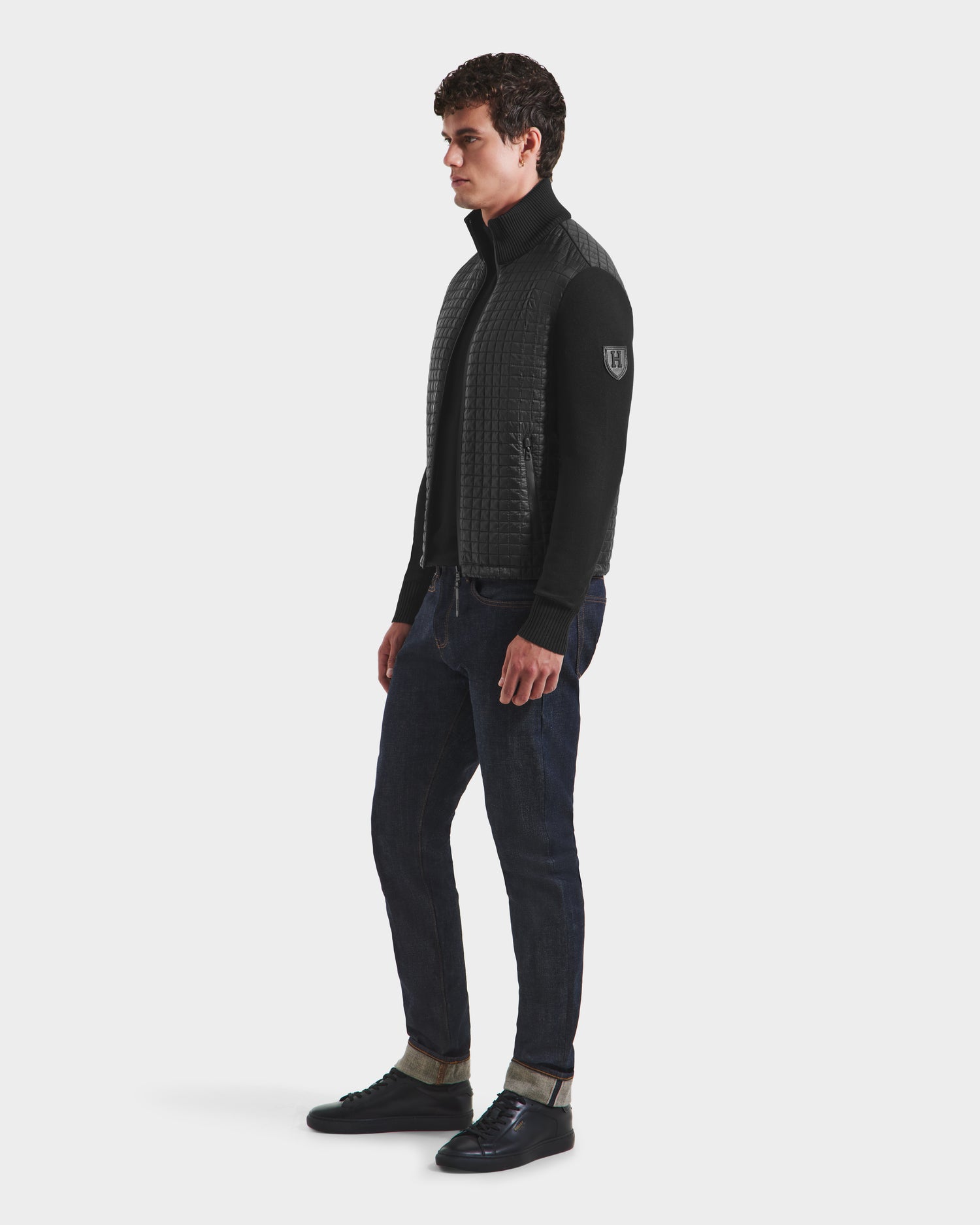 A man stands sideways against a plain white background wearing the Rudsak ABEL HV - H125501 Black men’s quilted hybrid puffer with ecofill insulation, paired with dark jeans and black sneakers.