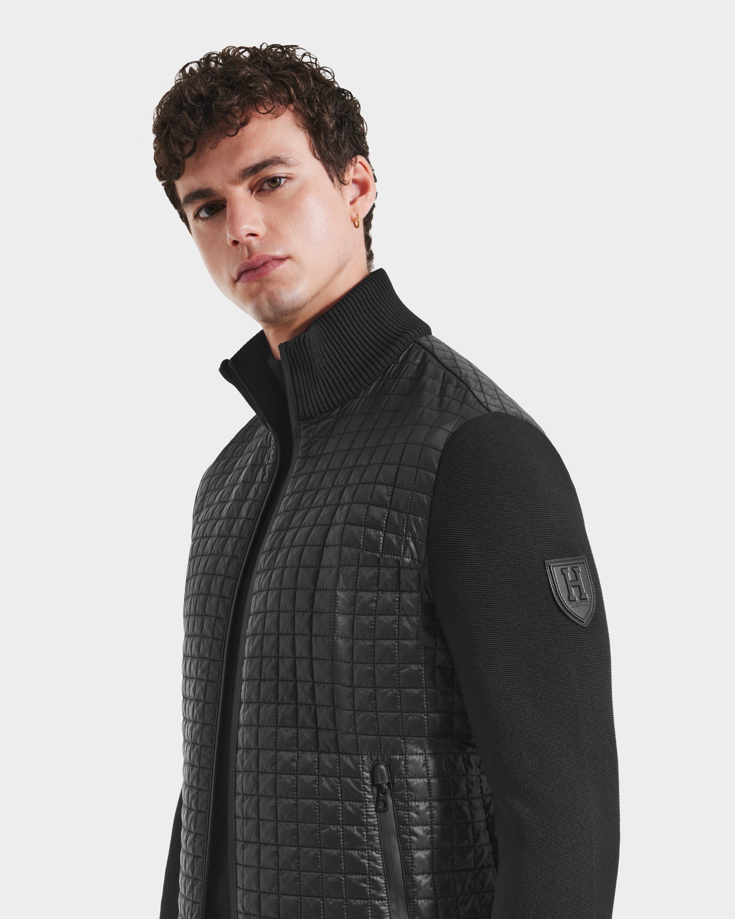 A young man with short, curly hair wears the Rudsak ABEL HV - H125501 Black men’s quilted hybrid puffer, featuring ecofill insulation, a high ribbed collar, zippered pocket, smooth sleeves, and a shield-shaped logo patch on the upper arm.