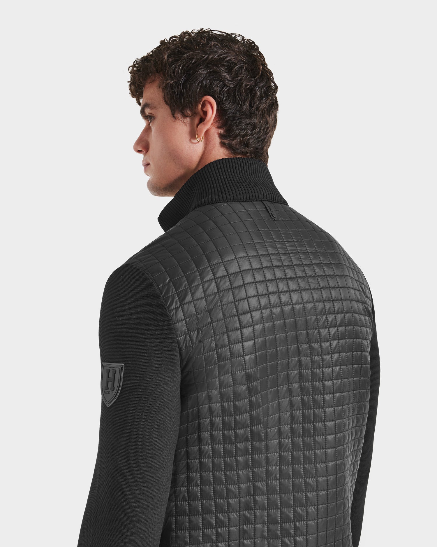 A man with short curly hair faces away, wearing the Rudsak ABEL HV - H125501 Black men’s quilted puffer with ecofill insulation, high ribbed collar, and H patch on the sleeve. The background is plain white.