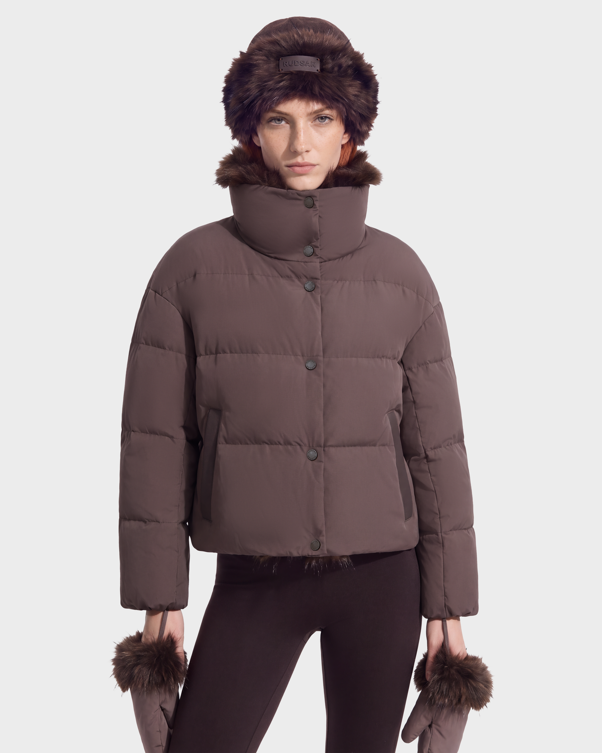 A model wearing the Rudsak ALONDRA - 8126992 Espresso dark brown cropped puffer jacket with a high collar, matching fur-trimmed hat, and matching mittens.