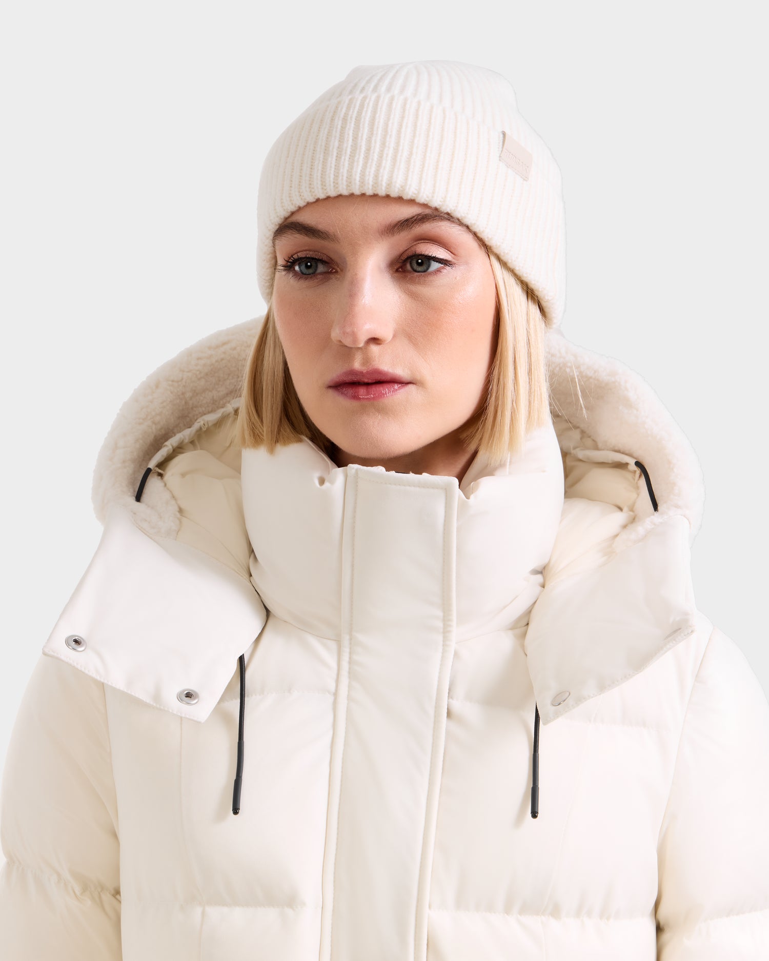 A woman with a neutral expression wears the Rudsak ANDREY - U425507 Vanilla unisex rib-knit beanie and a white puffer jacket, featuring a high collar and hood, standing against a plain light background—a true cold-weather essential.