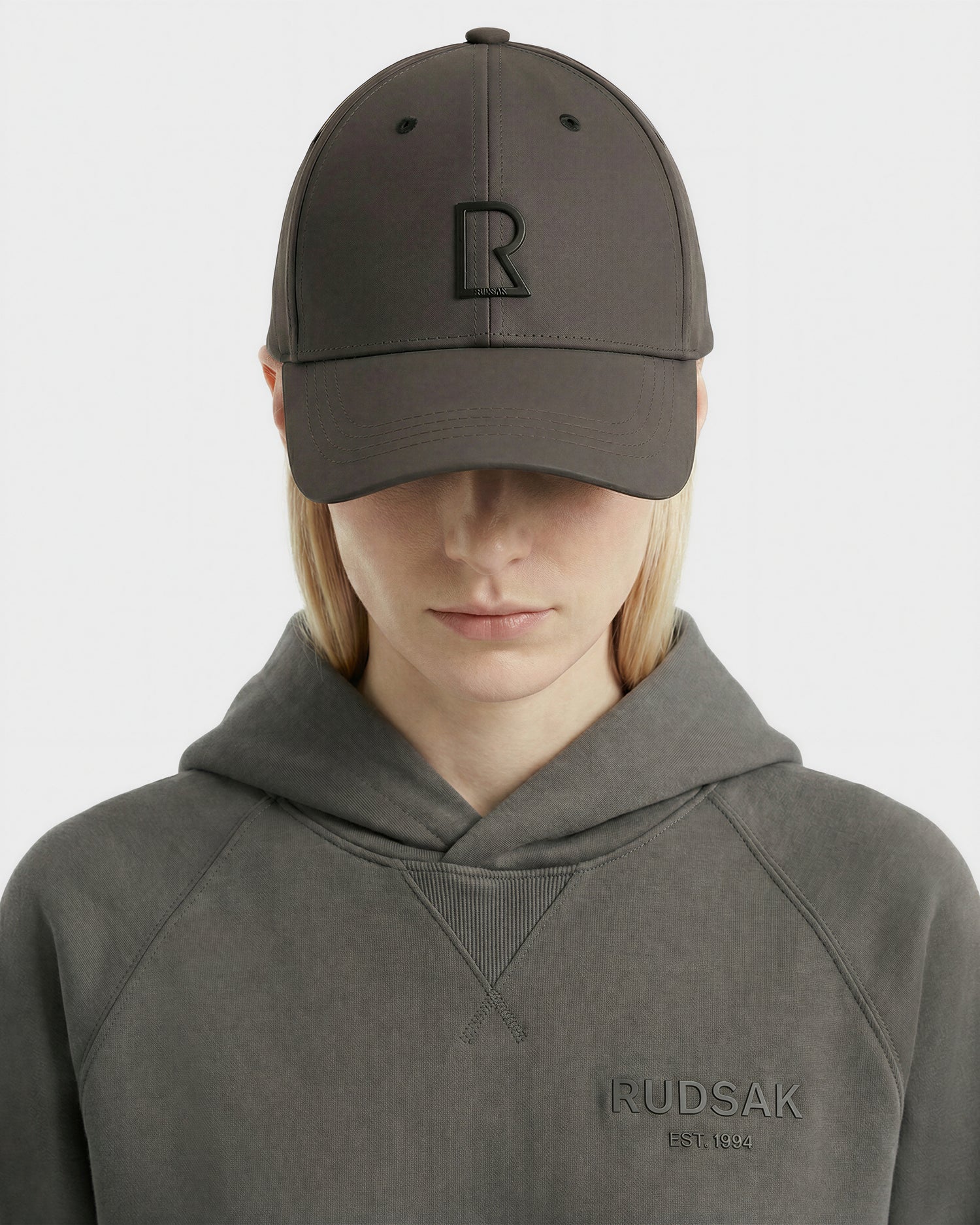 Wearing the grey Rudsak ARSENE - 9422504 unisex baseball cap with an embossed R logo and a matching grey sweatshirt, a person looks down against a plain white background.
