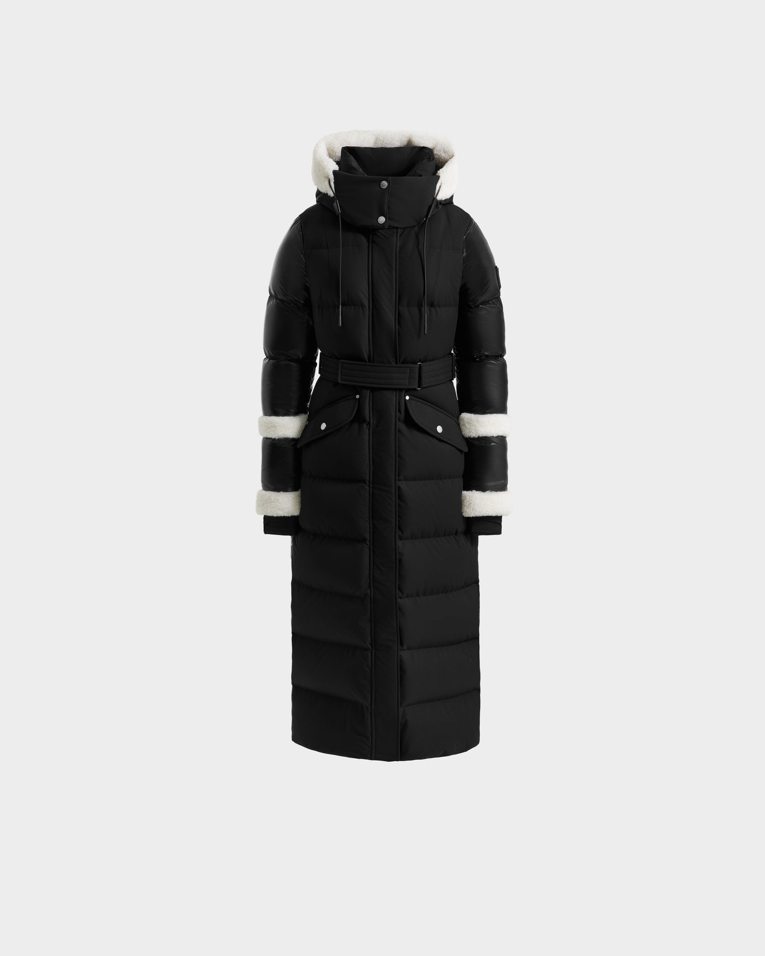 The AUDREY XL - 8125601 Black/Iceland by Rudsak is a maxi-length down puffer jacket with a belted waist, high collar, hood and cuffs lined in white faux fur, plus two front snap-closure pockets for winter warmth.