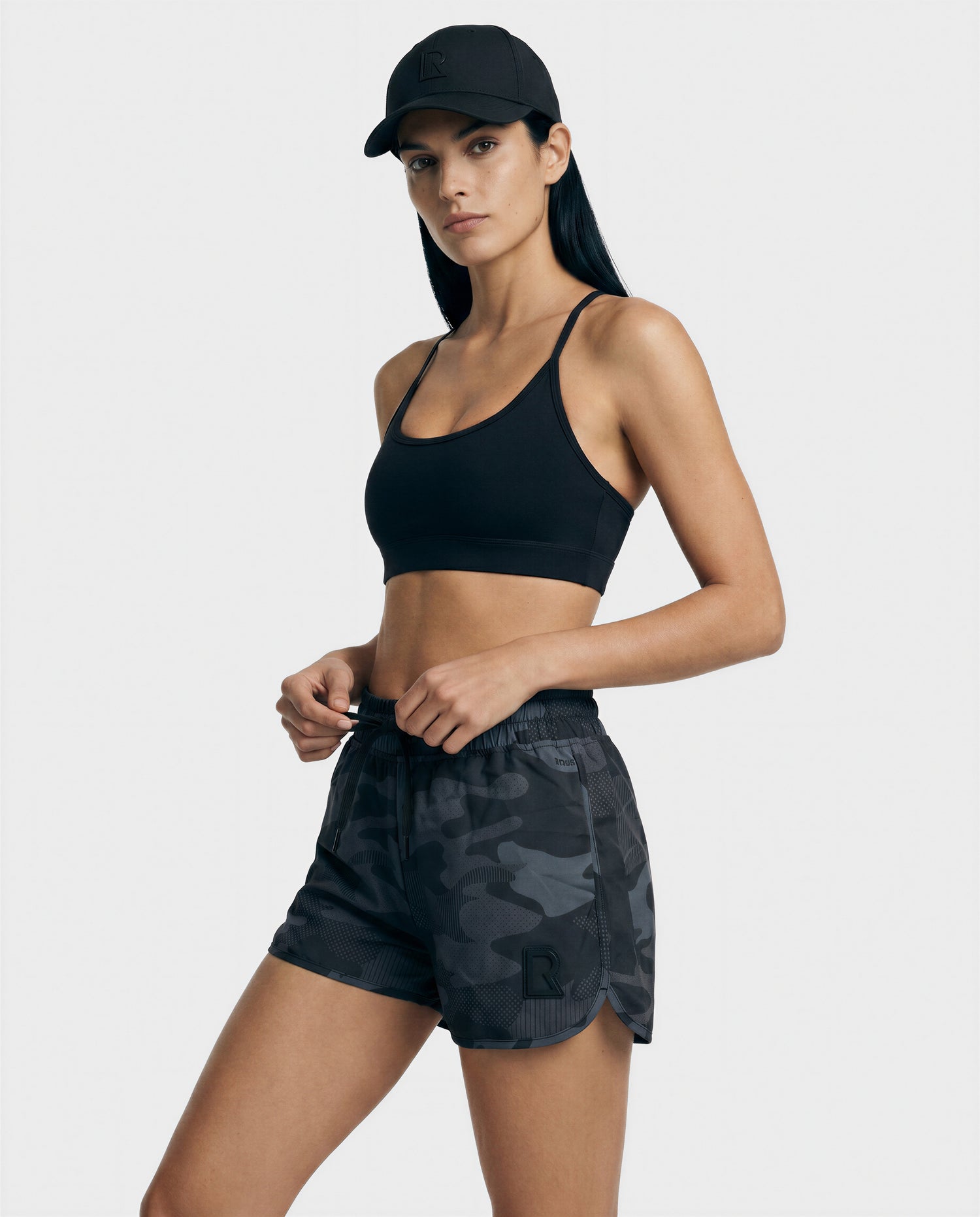 A woman wearing Rudsak's CAPRI - L523009 Black sports bra and black camouflage athletic shorts, paired with a black cap, stands against a plain light background, confidently facing the camera.
