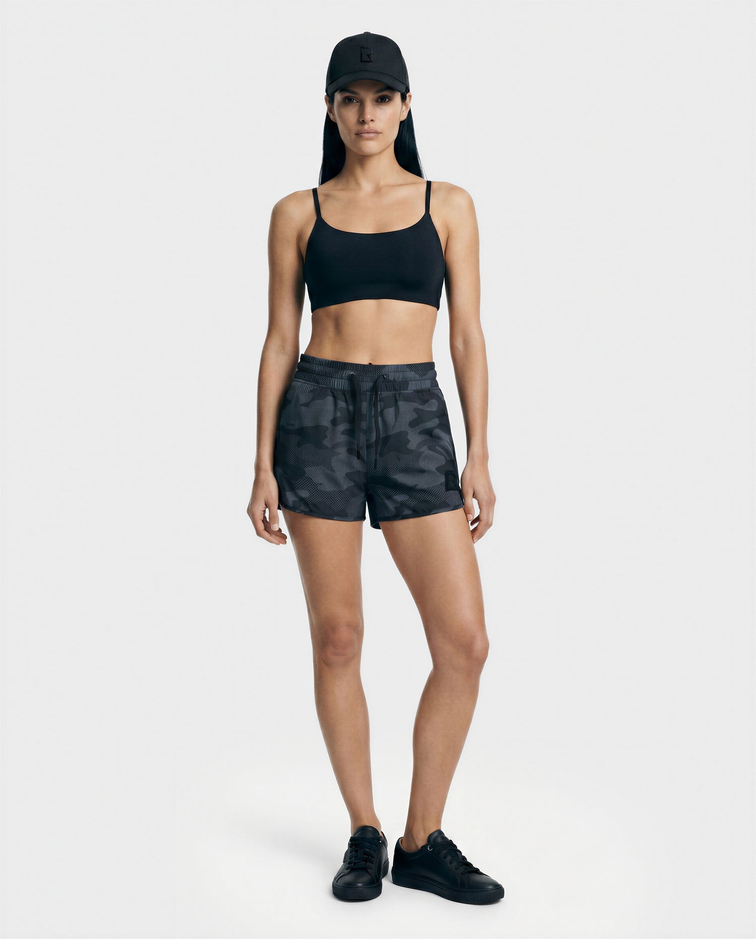 A woman faces forward on a plain light background, wearing Rudsak's CAPRI - L523009 Black sports bra and black camo athletic shorts, paired with black sneakers and a black baseball cap.