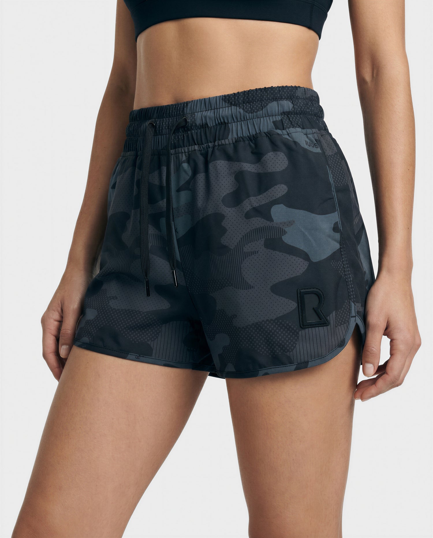 A person wears Rudsak's CAPRI - L523009 Black athletic shorts with an elastic waistband and a black 'R' logo on the left leg, paired with a black sports bra. Photo is cropped from mid-torso to mid-thigh.