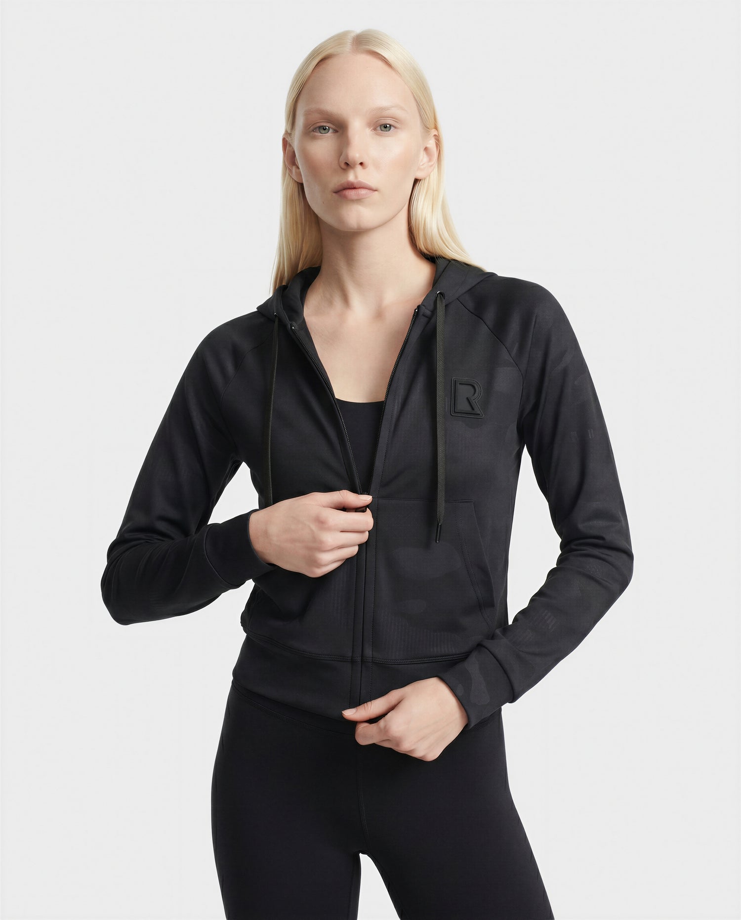 A woman with long blonde hair wears the Rudsak SAMMY - L523014T Camouflage Black top, standing against a plain white background and looking at the camera with a neutral expression.