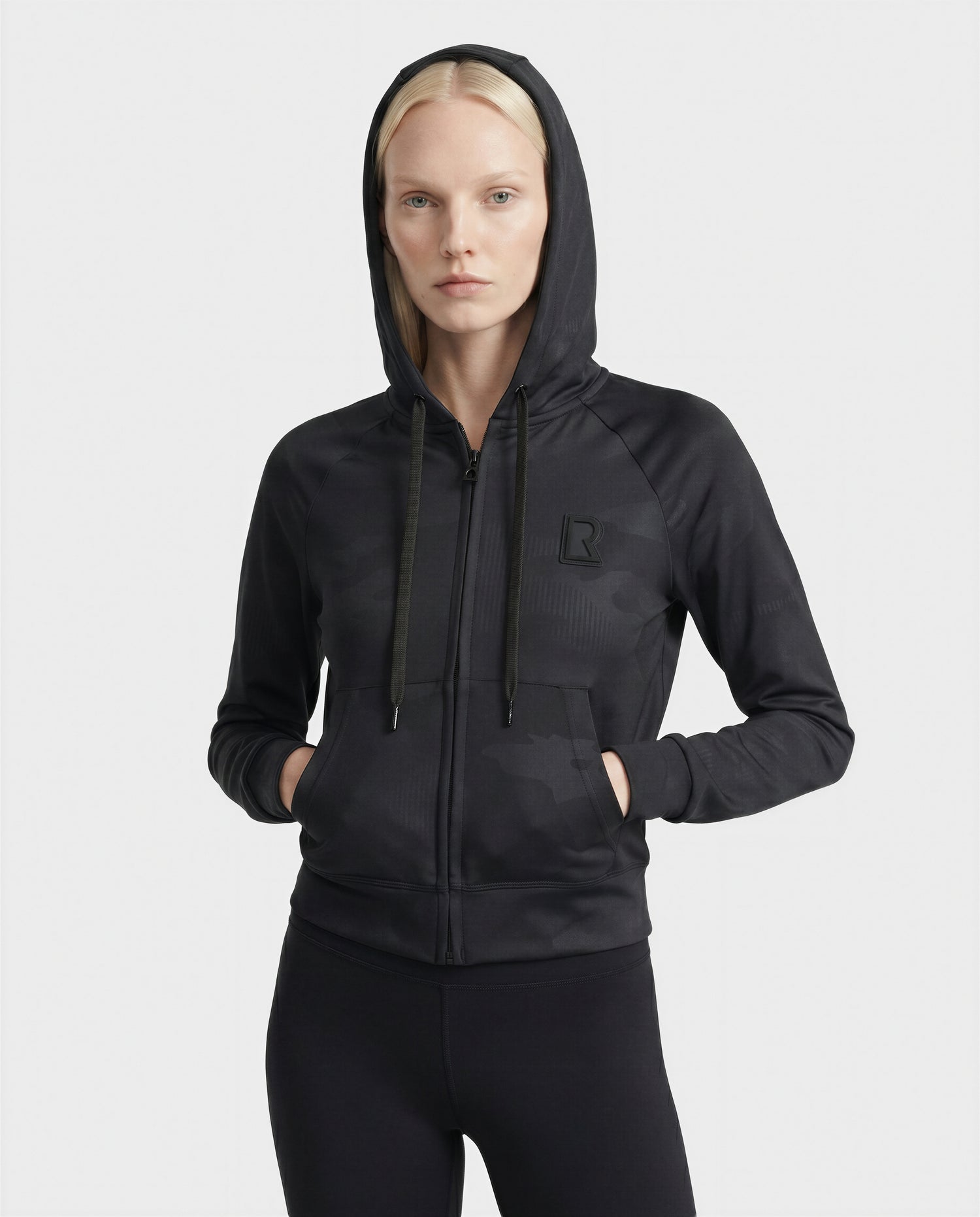 A woman with straight blonde hair wears the Rudsak SAMMY - L523014T camouflage black top, hood up and hands in her pockets, paired with black leggings against a plain white background.