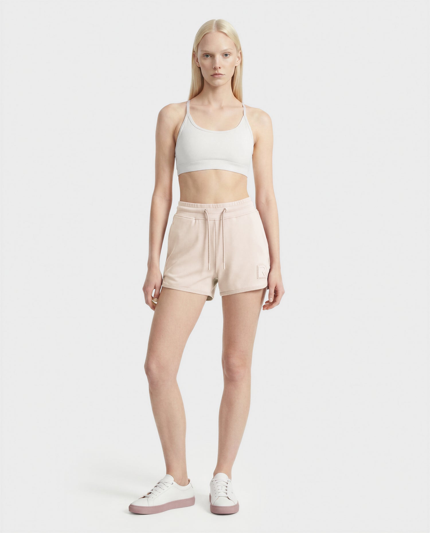 A woman with long blonde hair wears the Rudsak SAMMY - L523014B cream bottoms, a white sports bra, and white sneakers as she faces forward against a plain white background.