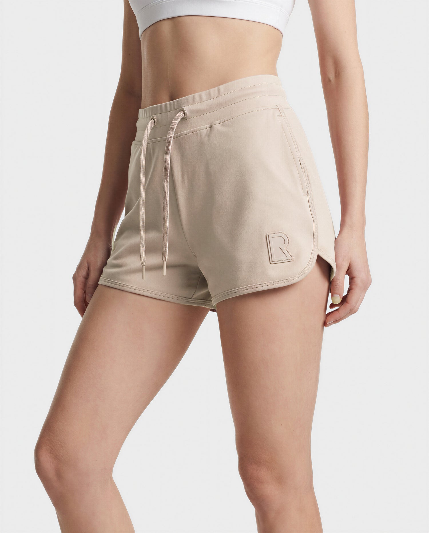 A person wearing the Rudsak SAMMY - L523014B cream shorts featuring a drawstring and raised R logo on the left leg, paired with a white sports bra, stands against a plain background.
