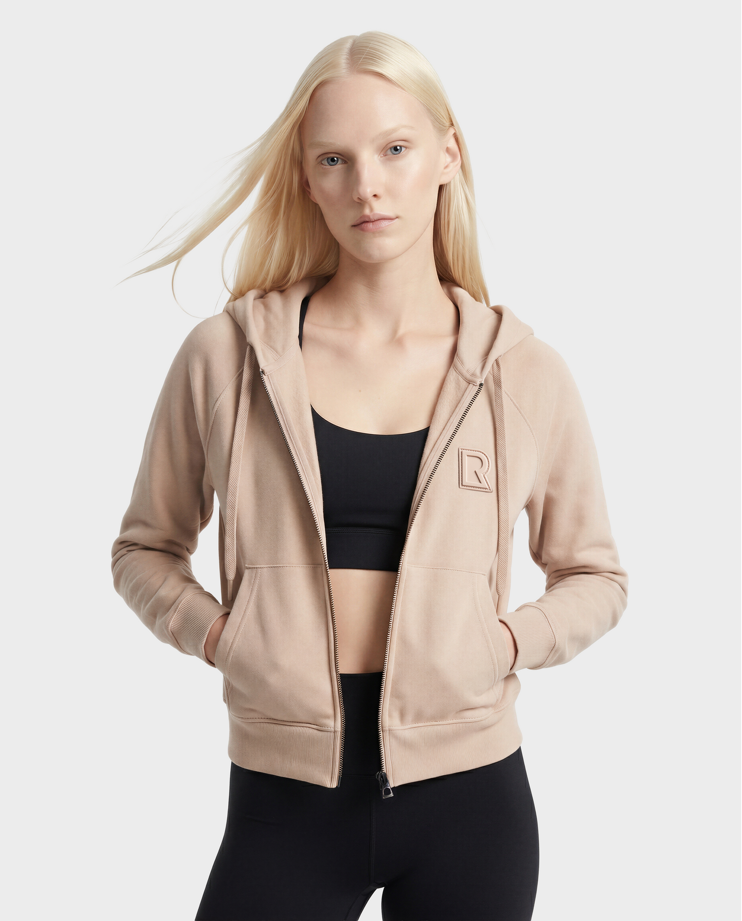 A woman with long blonde hair wears the Rudsak SAMMY - L523014T cream top, styled with hands in her pockets, against a plain light background.