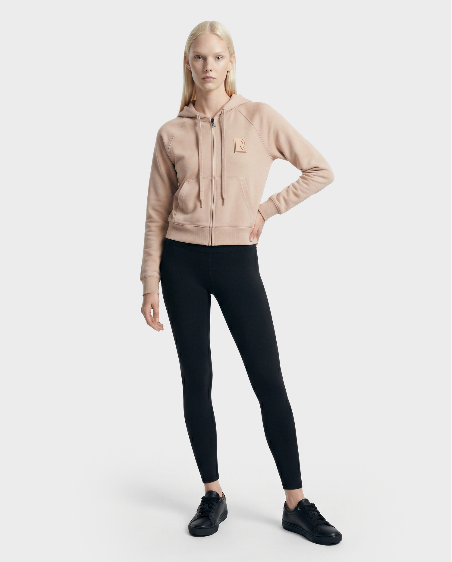 A woman with straight blonde hair and a neutral expression stands against a plain background, wearing the Rudsak SAMMY - L523014T Top in cream, black leggings, and black sneakers.