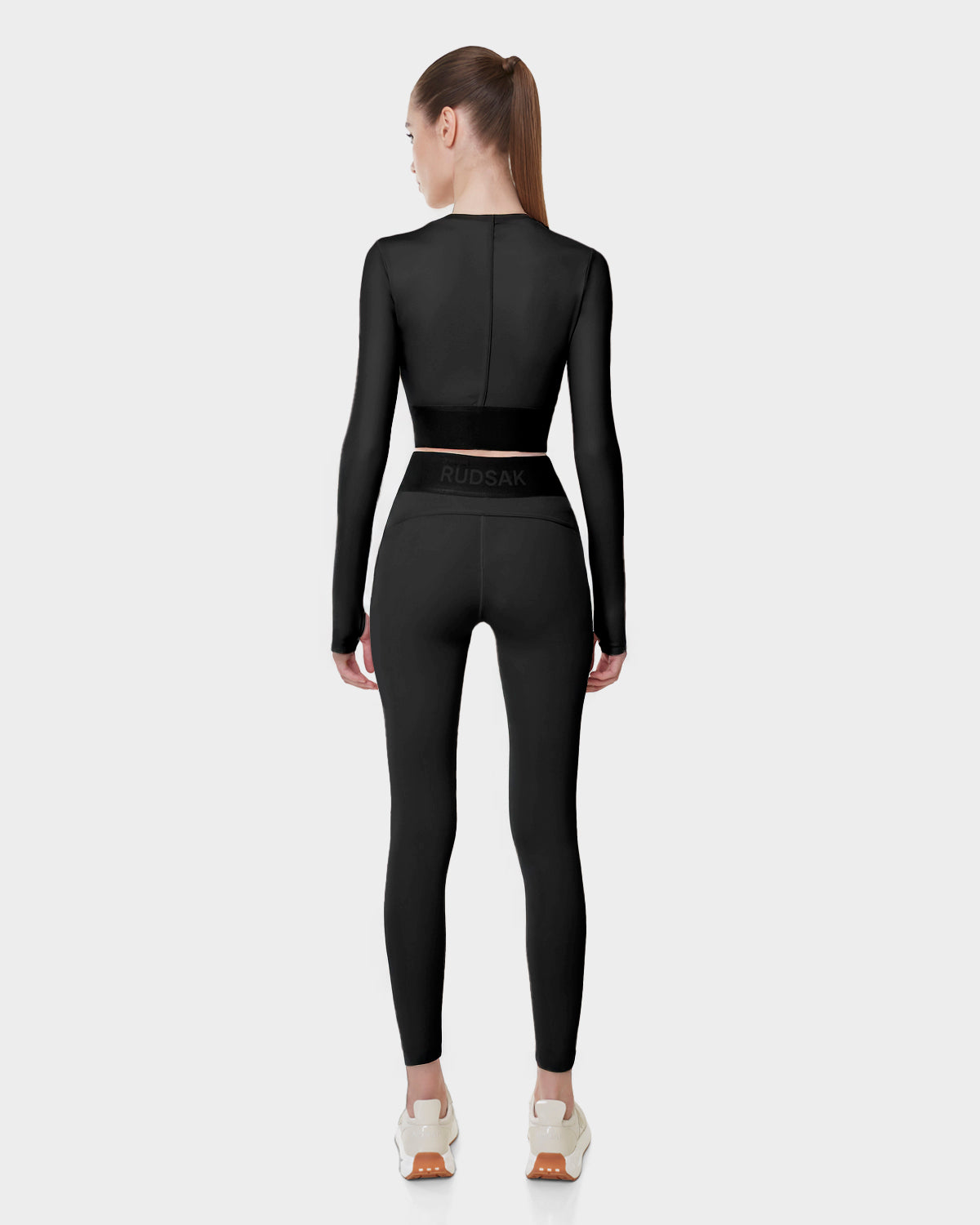 A woman with a ponytail faces away, showcasing athletic style in the Rudsak L523016T DOREEN - BLACK womens long sleeve sports top and matching leggings with branded waistband, paired with white sneakers, against a plain light gray background.