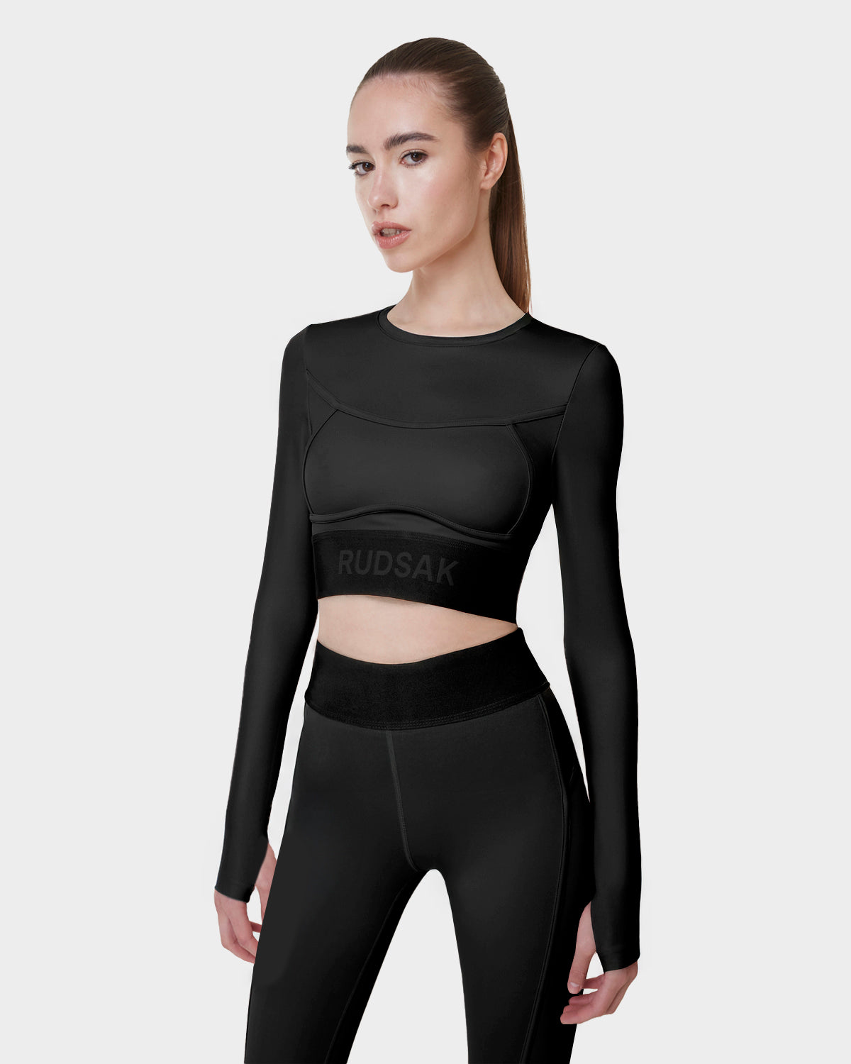 A woman with straight brown hair in a ponytail models the Rudsak L523016T DOREEN - BLACK fitted womens long sleeve sports top and high-waisted black leggings against a plain light grey background.