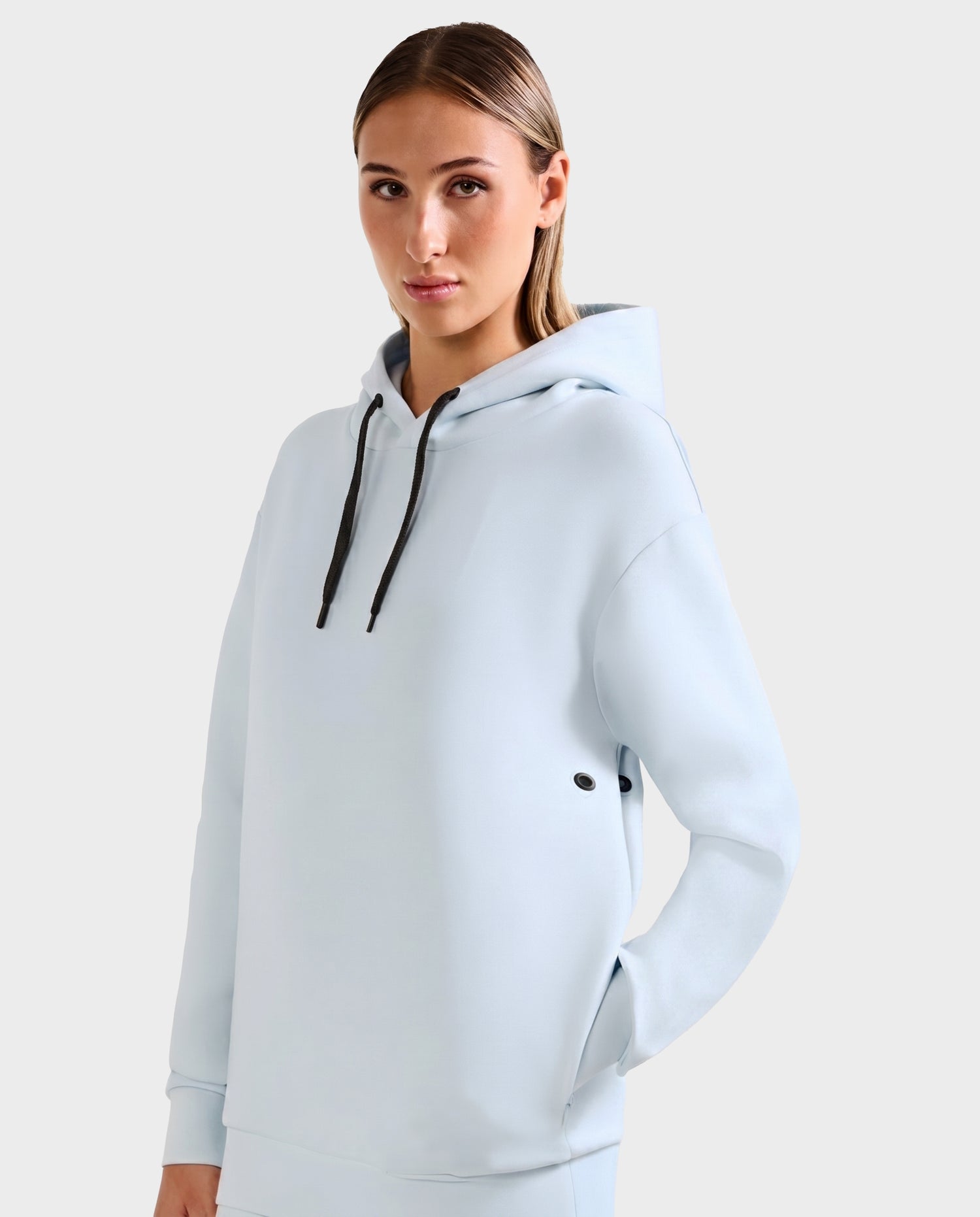 A woman with straight, light brown hair wears the Rudsak LEROY - U525509 Sky Blue W cotton-blend hoodie with black drawstrings, standing against a plain light gray background and looking directly at the camera with a neutral expression.