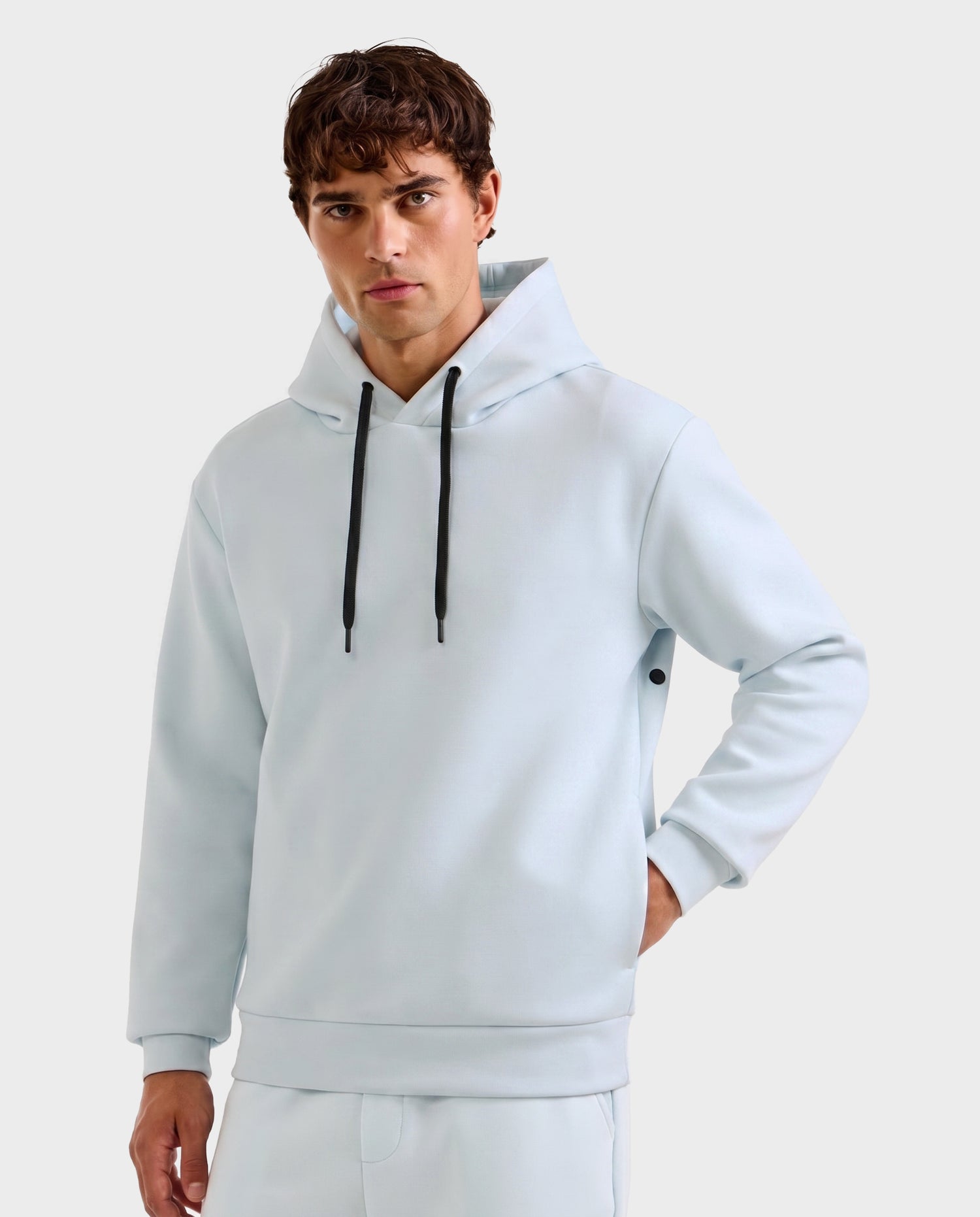 A young man with short brown hair models the Rudsak LEROY - U525509 Sky Blue M unisex hoodie and matching pants, featuring black drawstrings, as he stands against a light gray background with one hand in his pocket, gazing at the camera.