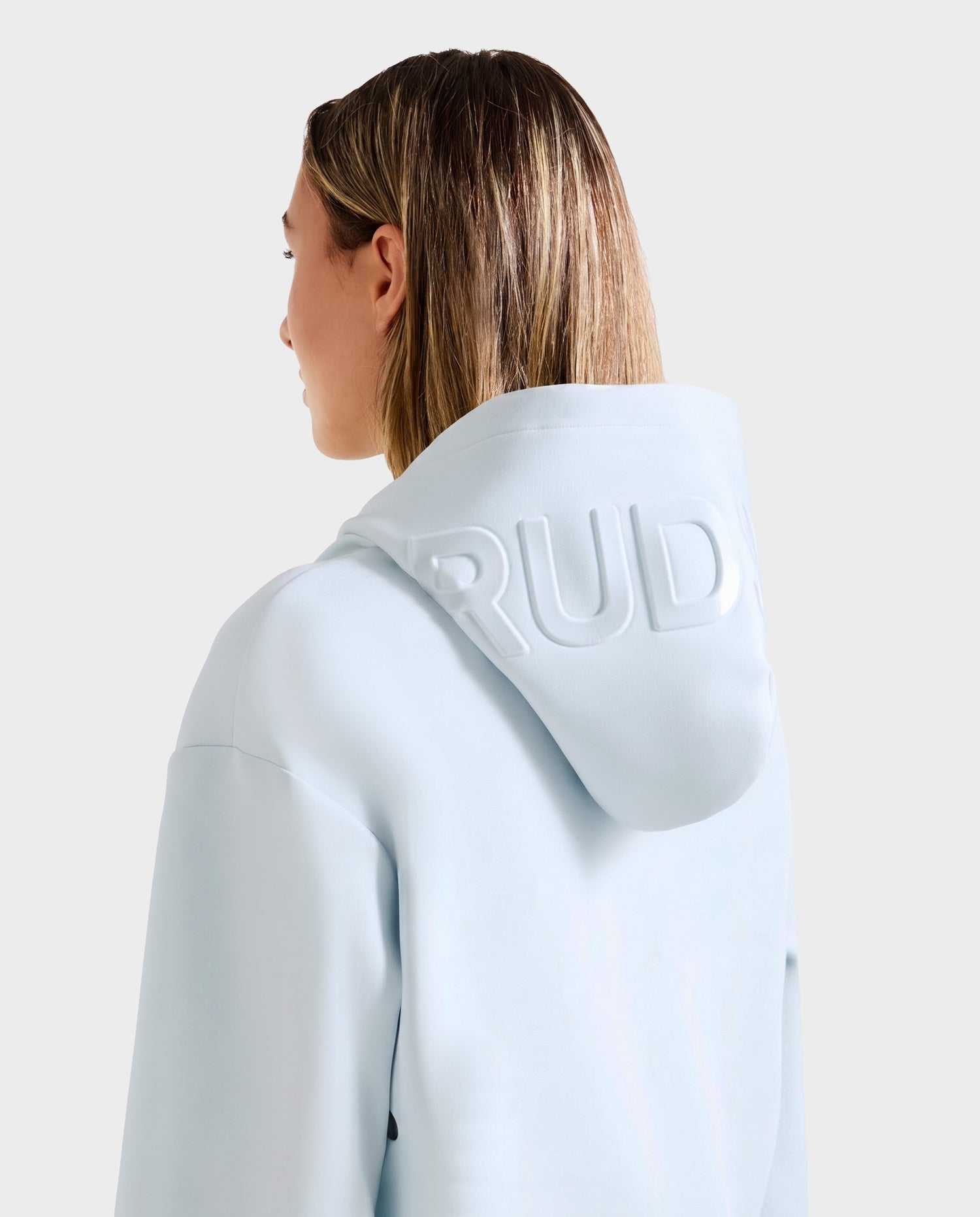 A person with straight, shoulder-length light brown hair is seen from behind wearing the Rudsak LEROY - U525509 Sky Blue W premium unisex hoodie, featuring "RUDY" embossed on the hood, against a plain light background.