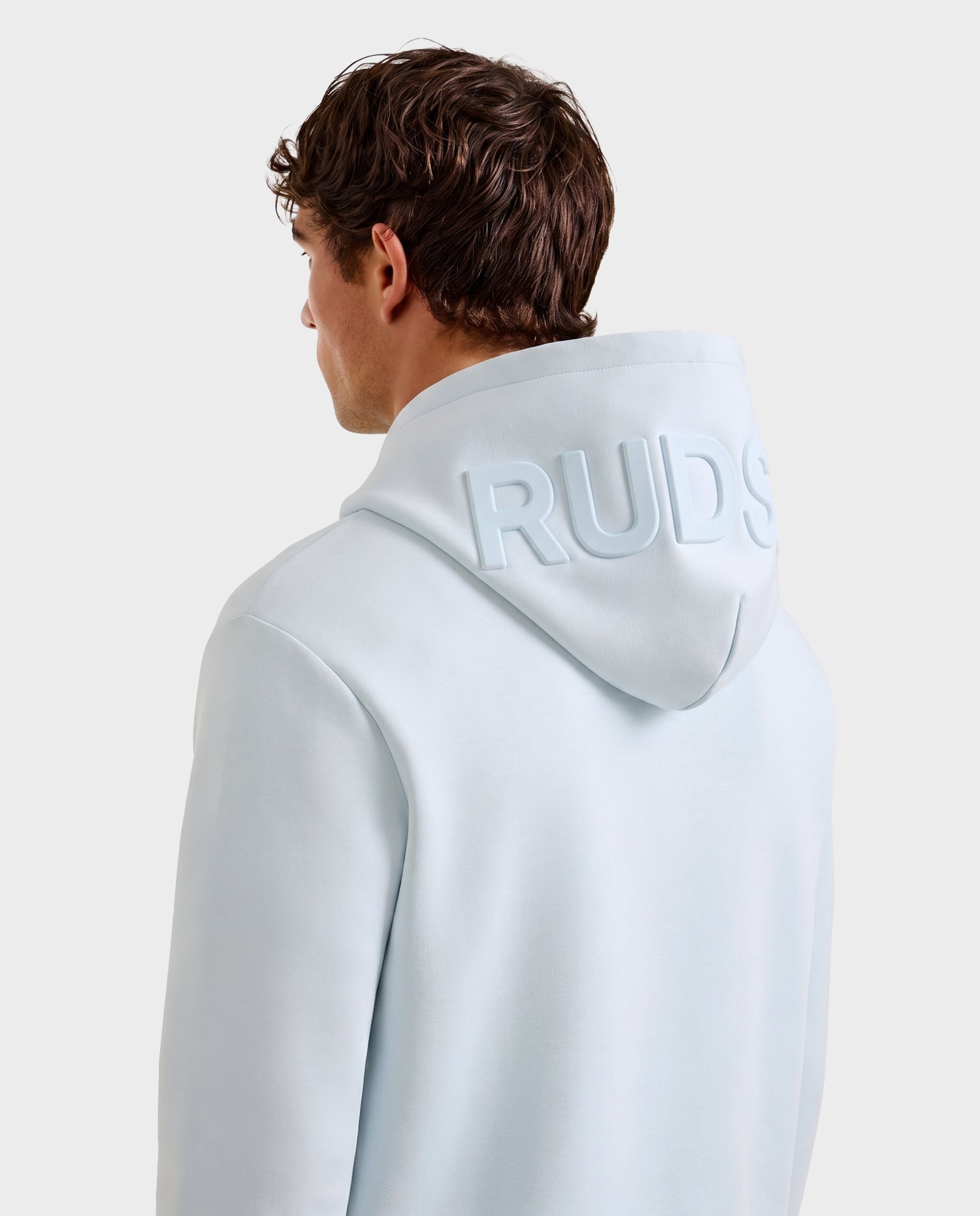 A person with short brown hair is seen from behind, wearing the Rudsak LEROY - U525509 Sky Blue M premium hoodie with "RUDS" embossed on the hood, set against a plain light gray background.