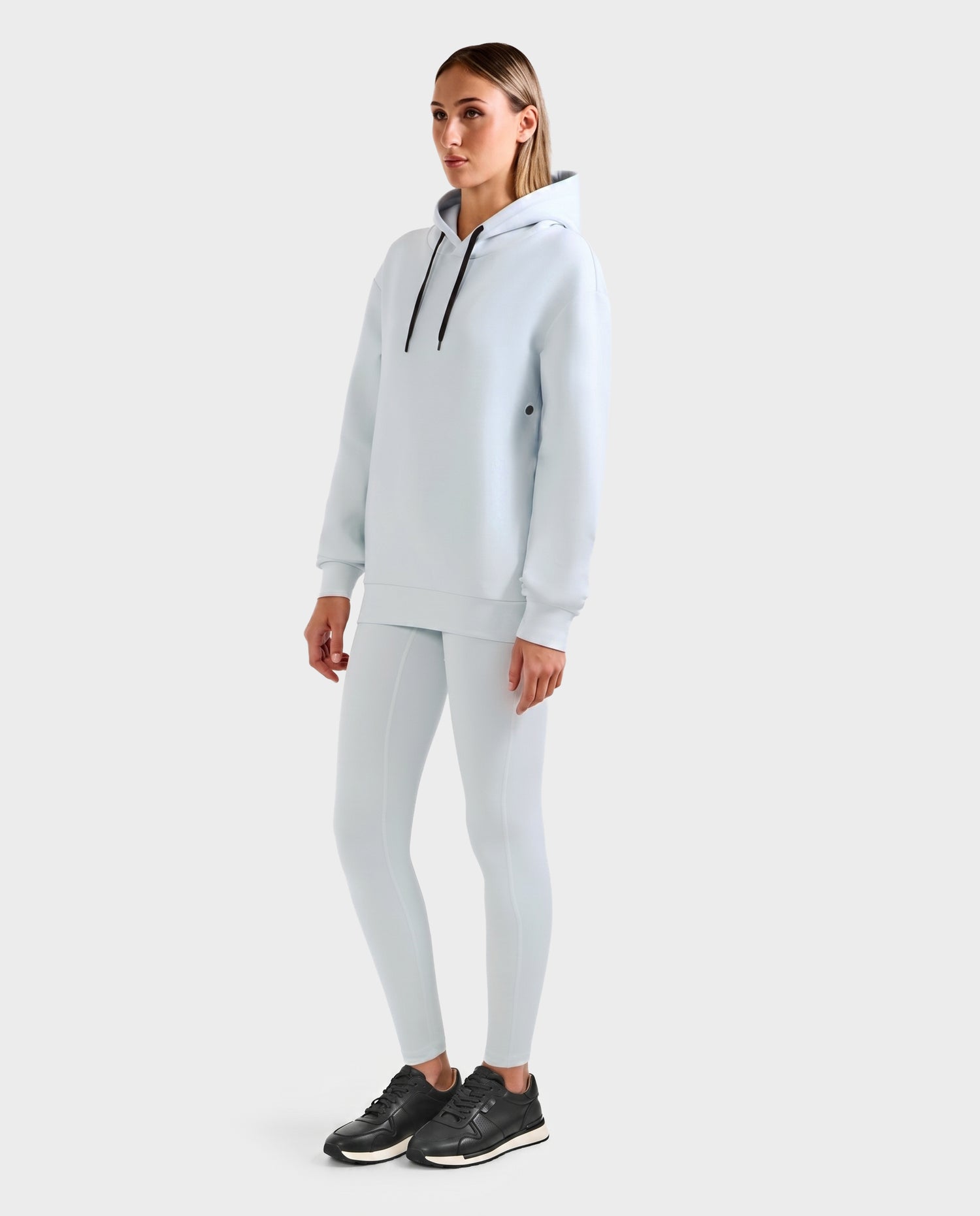 A woman stands against a plain background wearing the Rudsak LEROY - U525509 Sky Blue W hoodie and matching leggings, paired with black sneakers. She faces slightly to the side, arms relaxed at her sides.