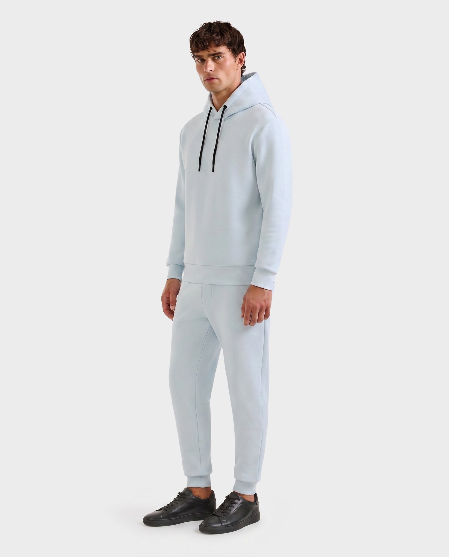 A man wears the Rudsak LEROY - U525509 Sky Blue M premium hoodie with black drawstrings and matching joggers, paired with black sneakers, set against a plain white background.