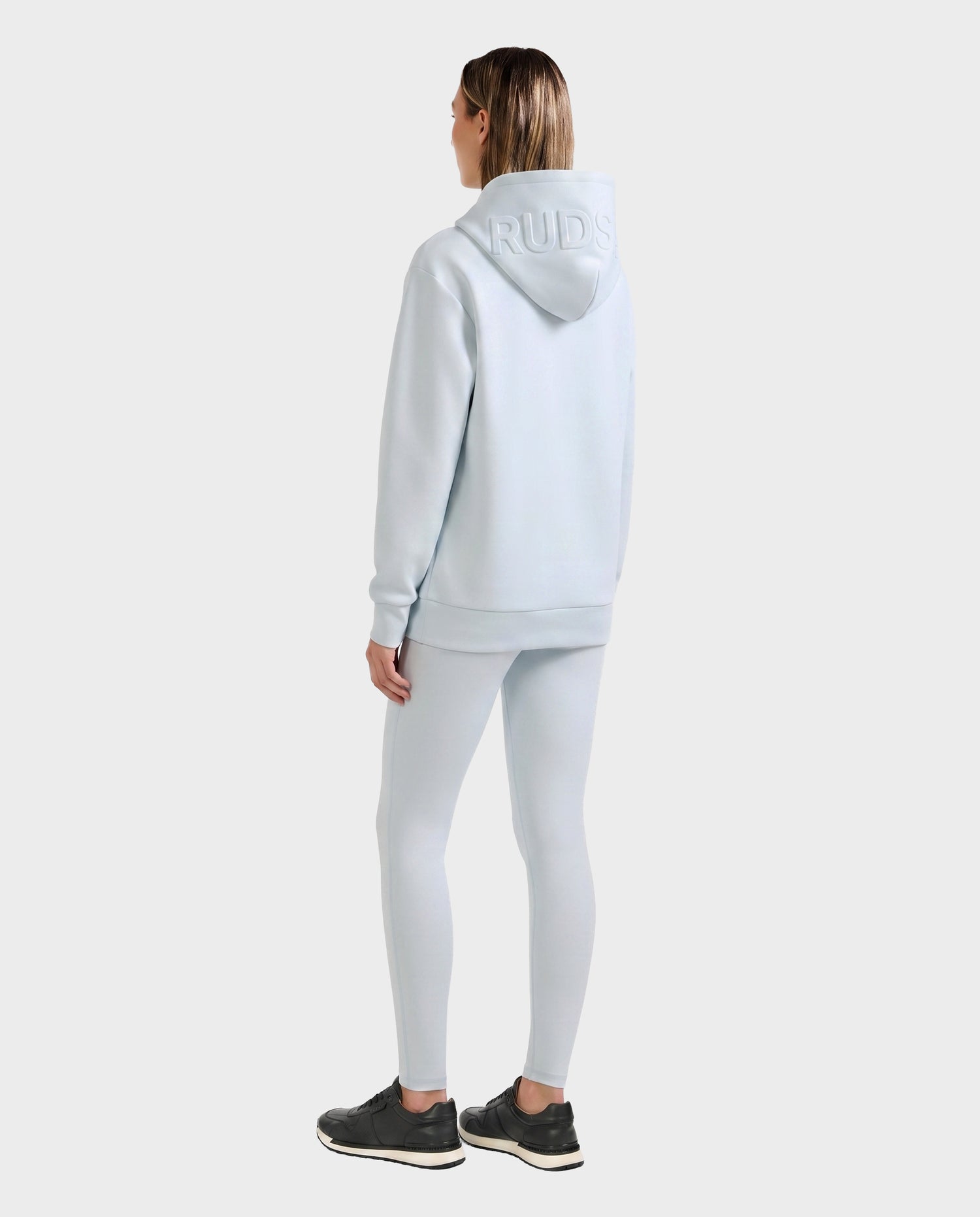 A person stands facing away against a plain light background, wearing the Rudsak LEROY - U525509 Sky Blue W hoodie with "RUDS" on the hood, matching leggings, and black sneakers, hands relaxed by their sides.