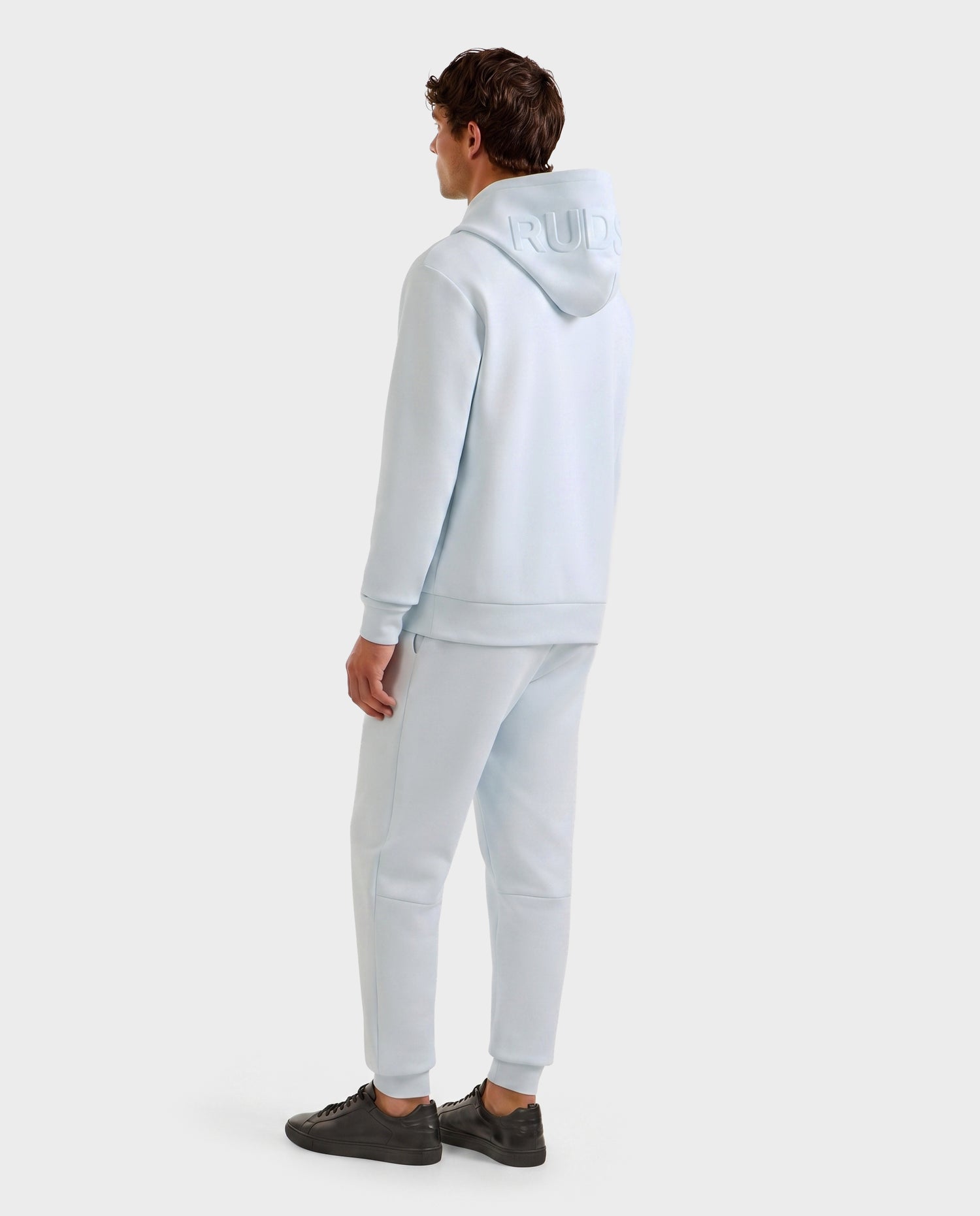 A person faces away, wearing Rudsak’s LEROY - U525509 Sky Blue M gender-inclusive hoodie and matching joggers with dark sneakers. The hoodie features raised text on the hood. The background is plain light gray.