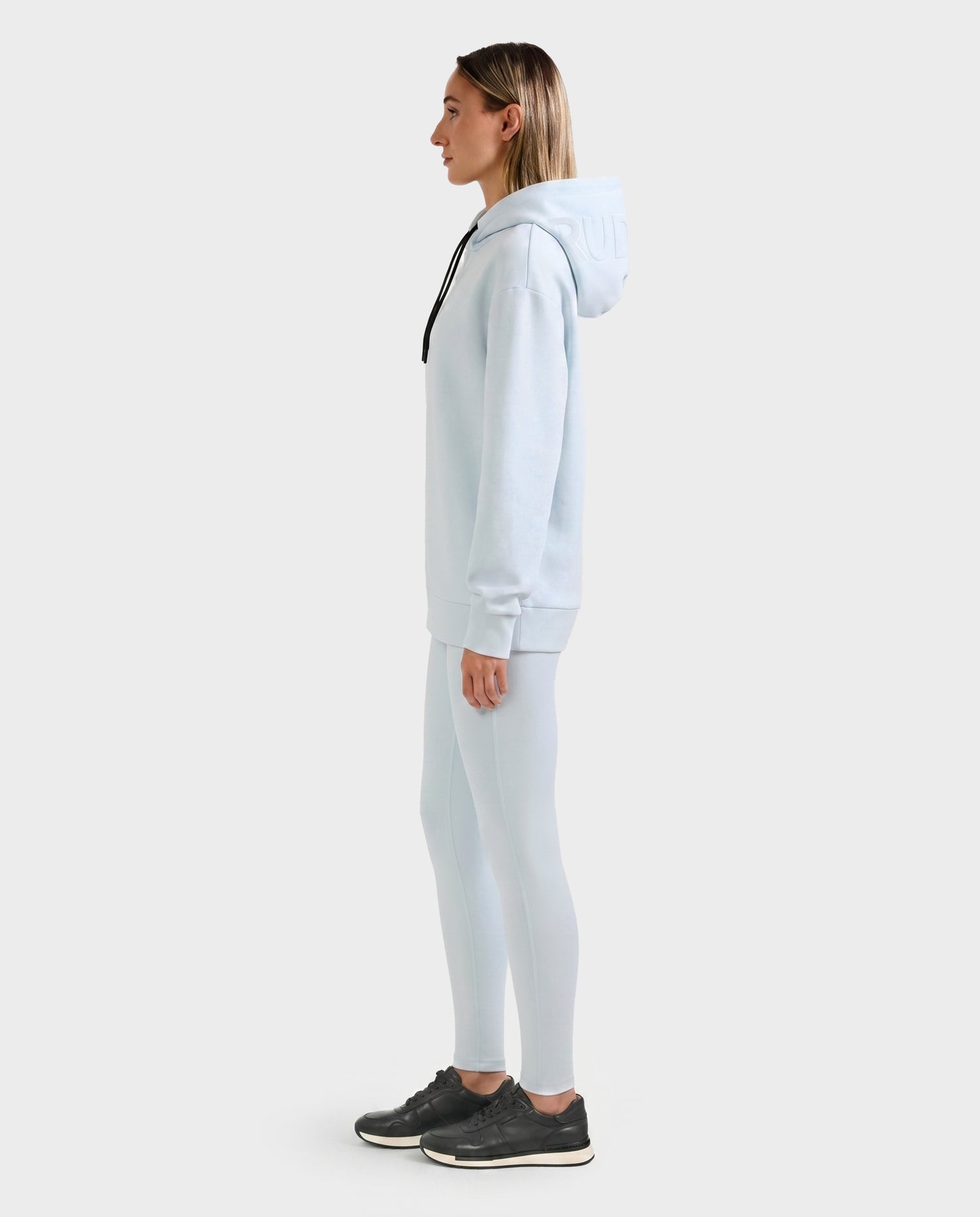 A person stands in profile wearing the Rudsak LEROY - U525509 Sky Blue W cotton-blend hoodie with matching leggings and black sneakers against a plain white background.