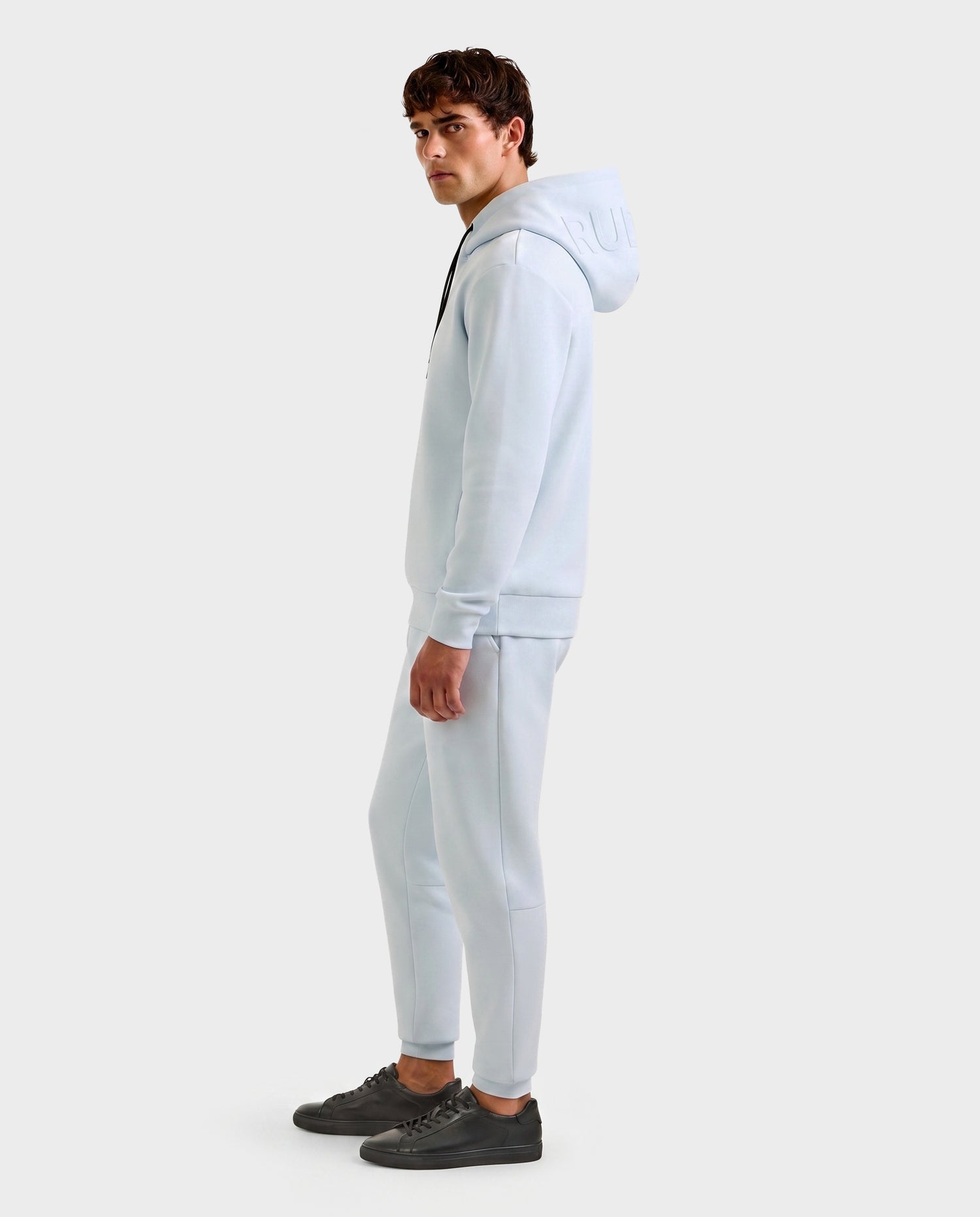 A man stands sideways against a plain light background, wearing the Rudsak LEROY - U525509 Sky Blue M premium hoodie and matching joggers, paired with black sneakers.