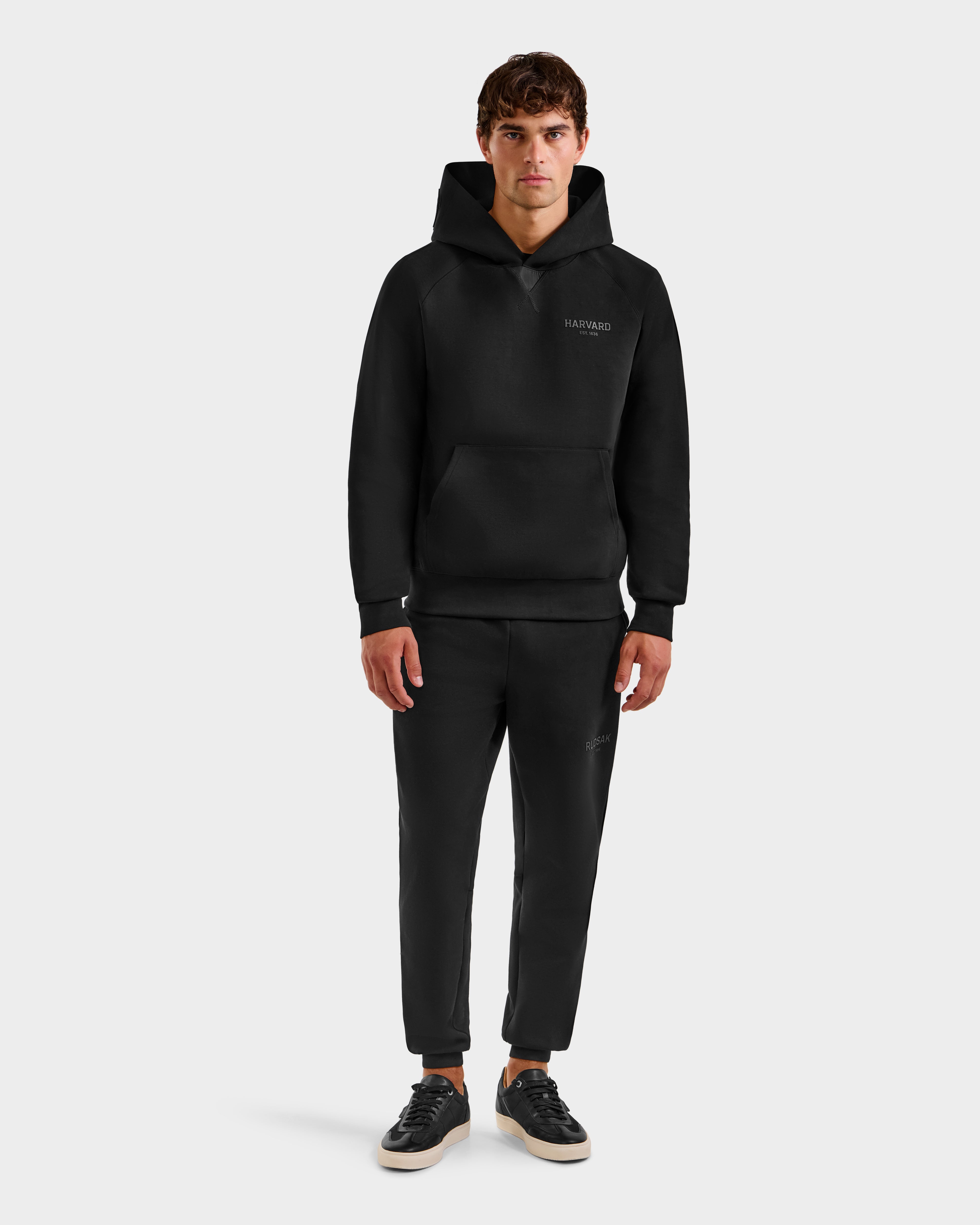 A person with short brown hair stands facing forward, wearing the limited-edition Rudsak BARRY HV - H525500 Black hoodie and matching black jogger pants, paired with black sneakers featuring white soles. The background is light gray.