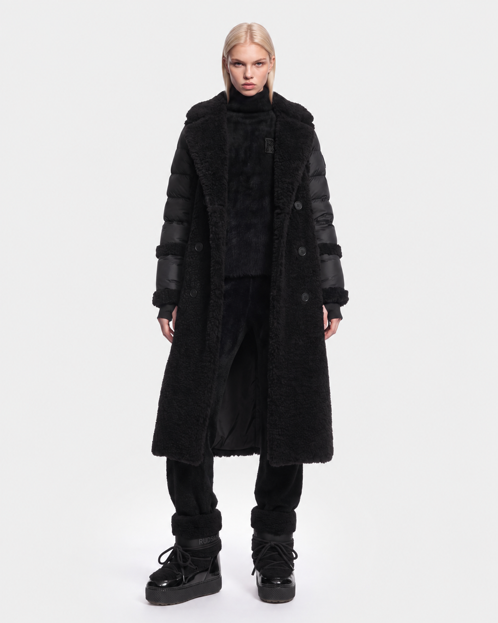 A person with straight blond hair wears the Rudsak NOELLE - 8126971 Black long hooded coat, black pants, and black platform boots, standing against a plain white background.