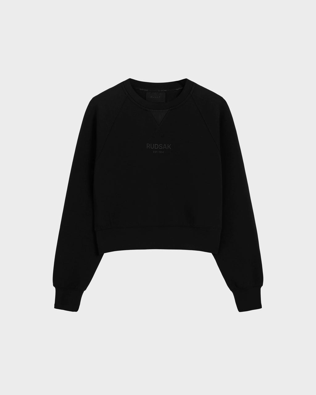 The BETSY - 8525087 Black is a black crewneck sweater by Rudsak with ribbed cuffs and hem, featuring the brand subtly printed on the front in a modern cropped silhouette, shown against a plain light grey background.